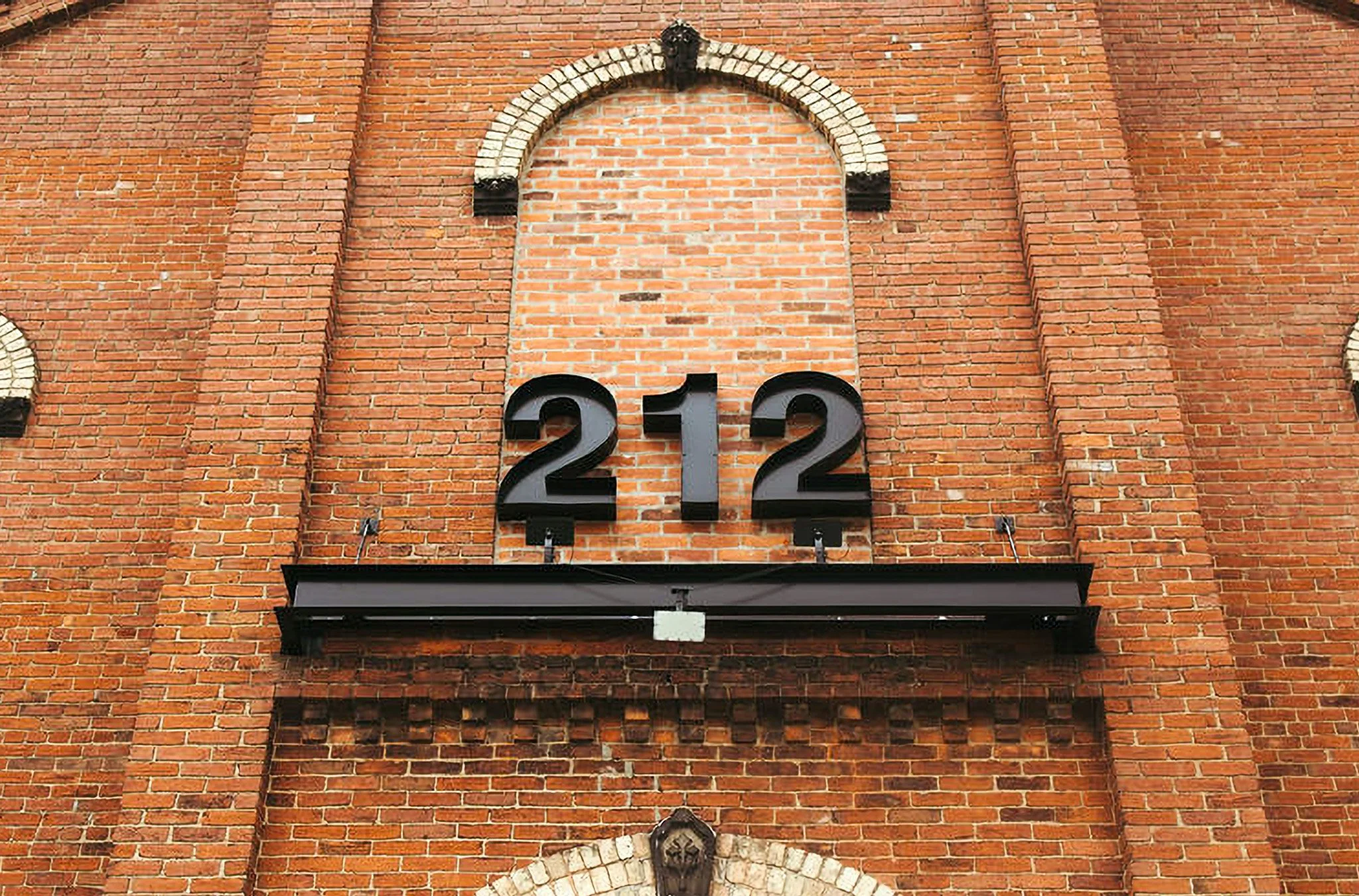Close-up of a brick building facade with black metal numbers '122' mounted below a canopy, and an arched brick decorative element above.