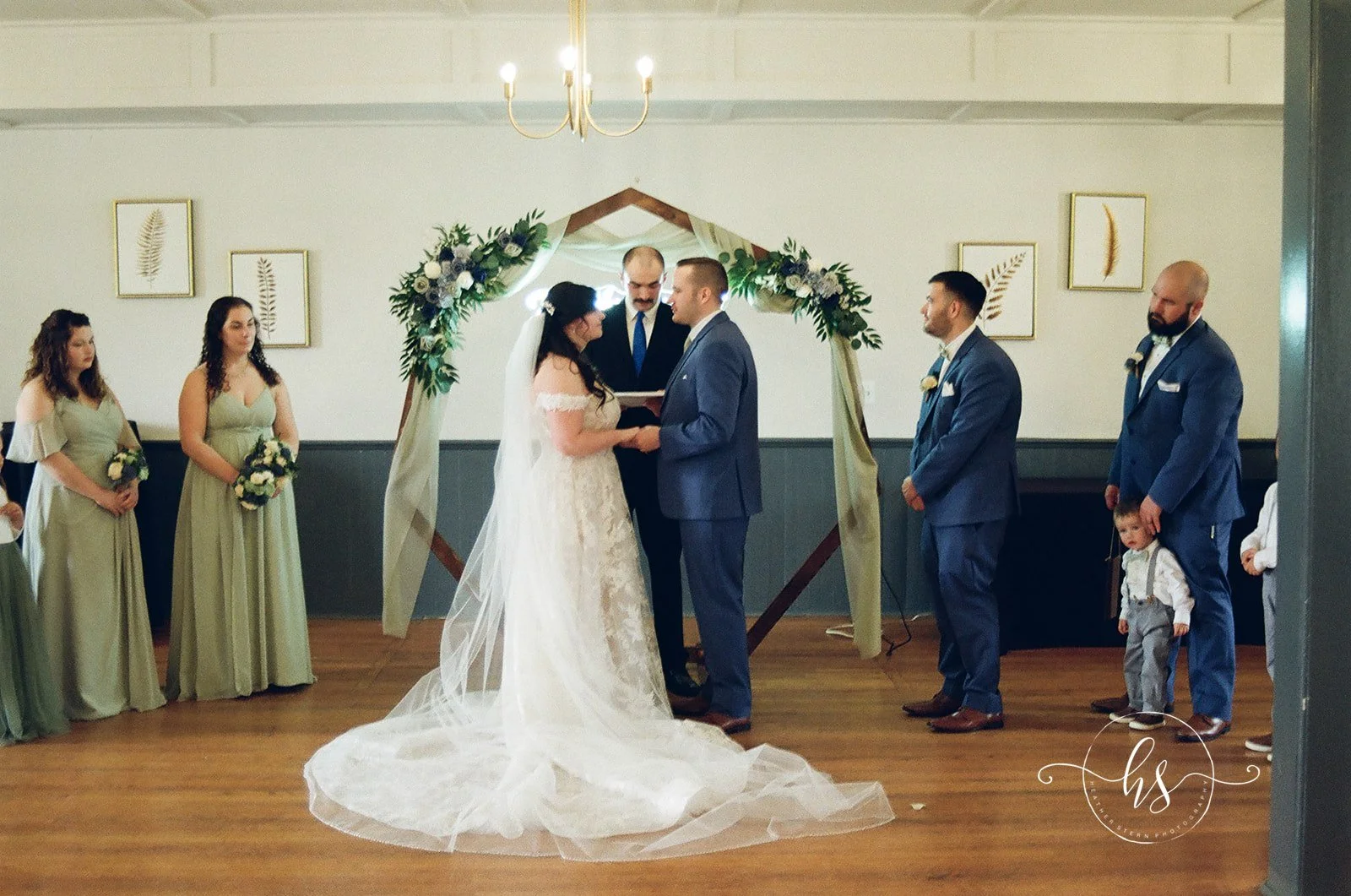 A couple exchanges vows during their wedding ceremony inside a decorated hall. The bride is wearing a lace wedding gown and veil, and the groom is in a blue suit. Bridesmaids and groomsmen stand nearby, and an officiant conducts the ceremony under a 