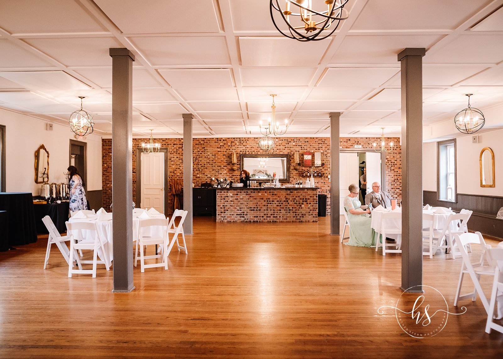 Wedding reception area with round tables covered in white tablecloths and white chairs. A brick fireplace wall with a mirror, a bartender behind a brick bar, and guests seated at a table. The room features wooden floors, decorative light fixtures, an