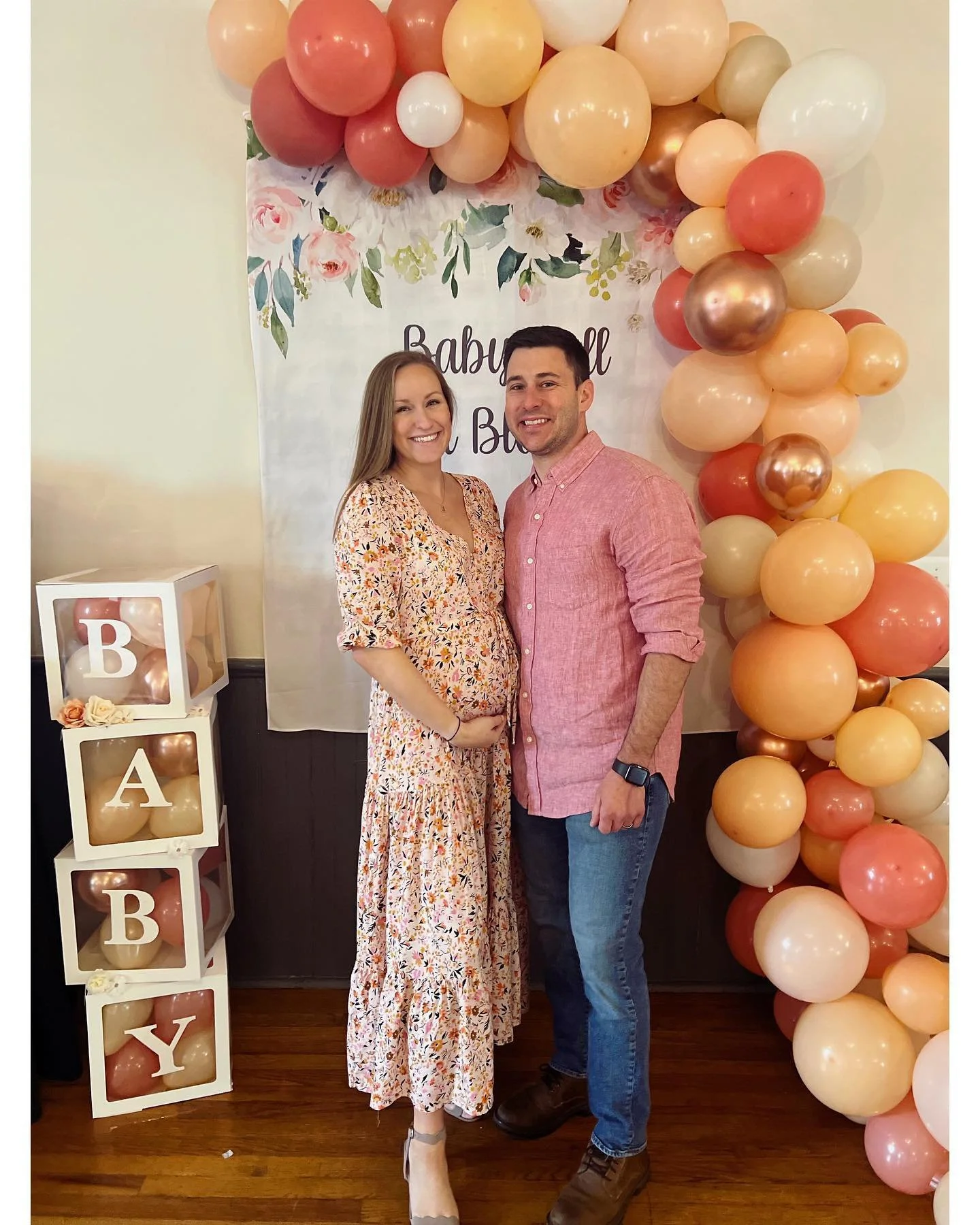 A pregnant woman and a man standing together at a gender reveal party, smiling. Behind them is a backdrop with pink and peach balloons and floral decorations that reads 'Baby Girl'. To the side, a white cube display spells out 'BABY' with balloons in