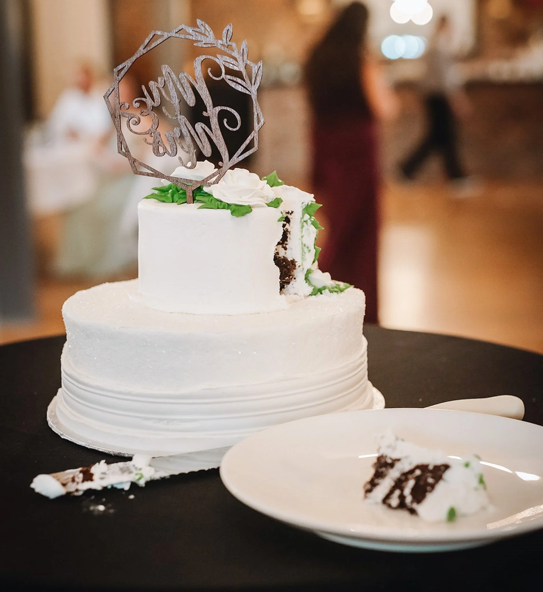 A slice of cake with white frosting, green decorative leaves, and a cake topper with script text surrounded by leaves, on a black table with crumbs and partial plates visible, with a blurred person in background.