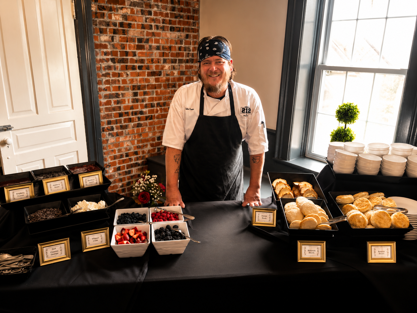 A smiling man in a chef uniform and apron stands behind a dessert table with berries, strawberries, blackberries, and various baked goods, with a brick wall and large window in the background.