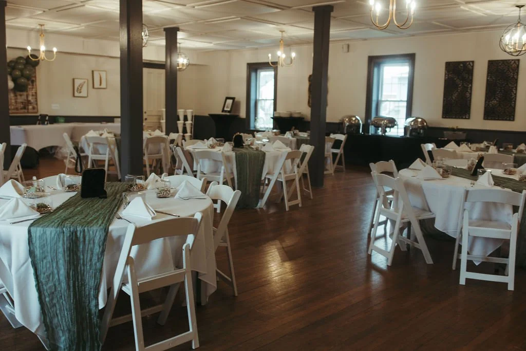Dining room decorated for an event with round tables covered in white tablecloths, green table runners, and neatly folded napkins; chairs around each table; chandeliers hanging from the ceiling; windows letting in natural light; wall art and balloons