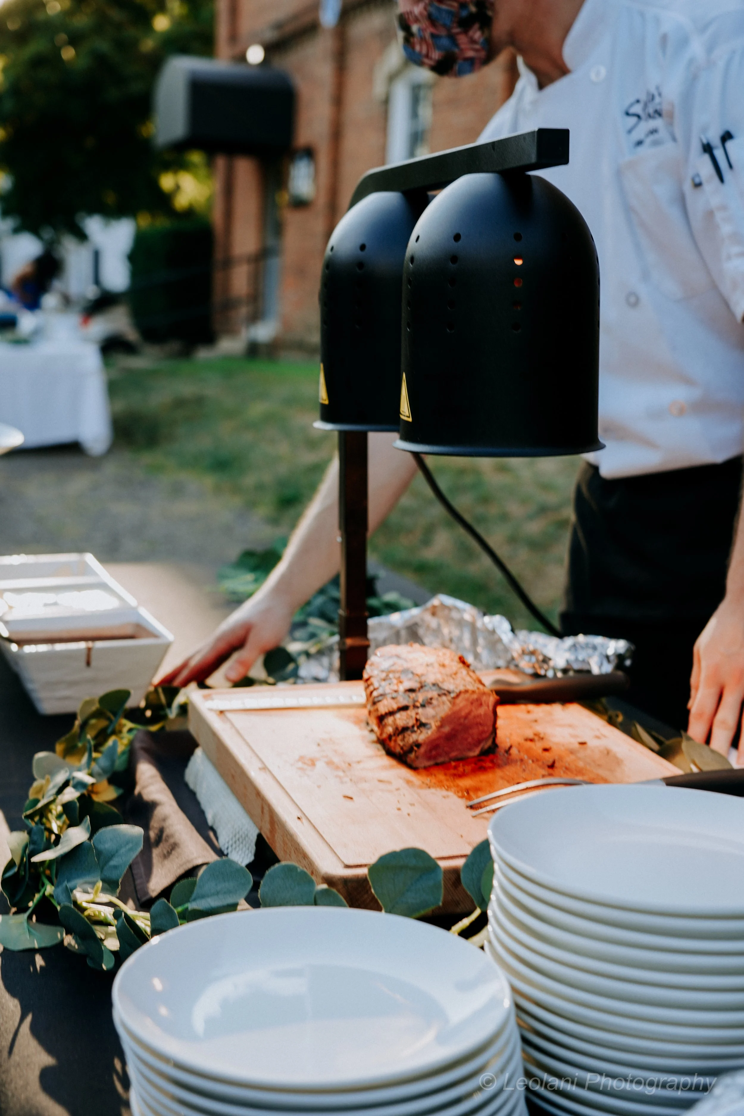 A person in a white chef's coat and colorful headscarf is preparing food outdoors. There are two large black sous-vide machines on a stand, and slices of cooked meat on a wooden cutting board. Stacked white bowls are in the foreground, with additiona
