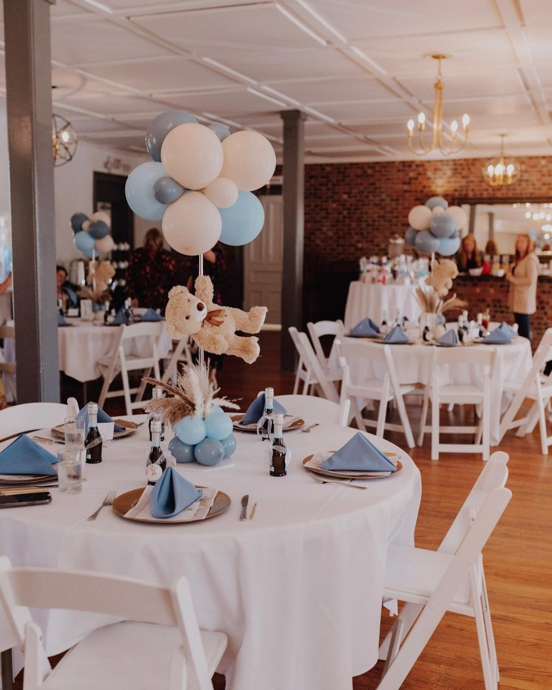 Decorated banquet room with round tables set for a celebration, featuring blue, white, and gray balloon centerpieces with teddy bear plush toys, blue napkins, and tableware on white tablecloths.