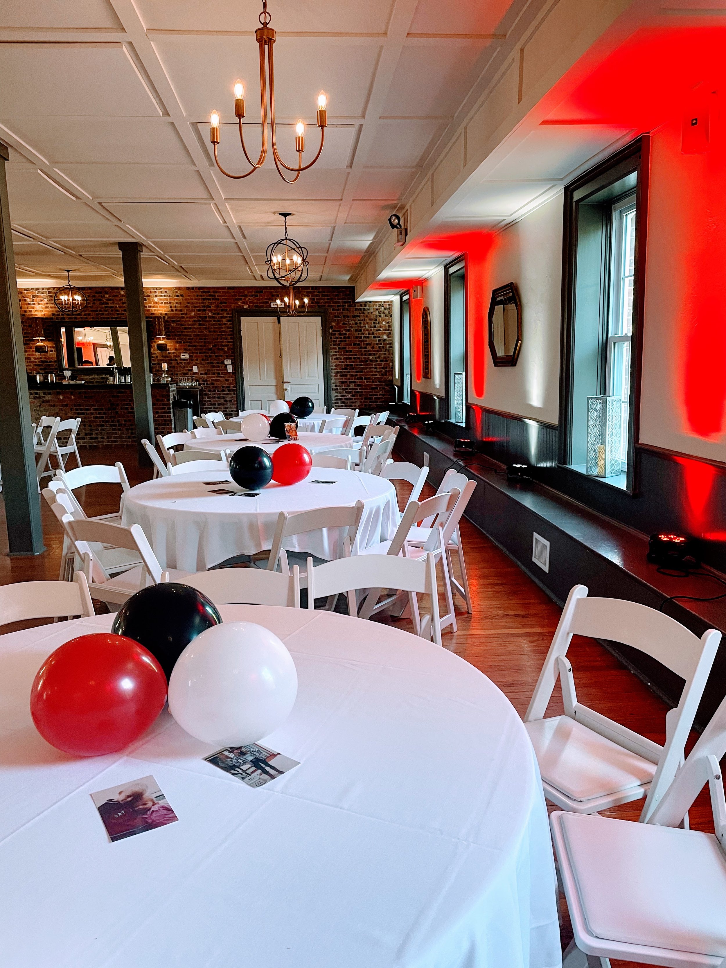 Round white tables decorated with black, white, and red balloons, with chairs arranged around them, set in a venue with brick walls, large windows, and decorative ceiling lights for a celebration or event.