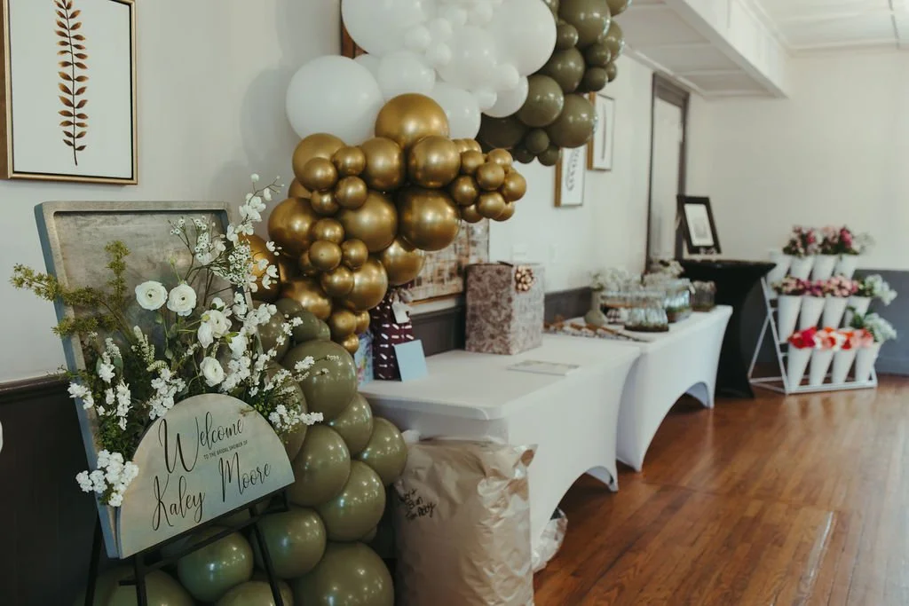 Decorative event setup with balloon arrangement including white, gold, and gray balloons, a floral display with white flowers and greenery, a welcome sign for Kaley Moore, and a table with party favors and decorations in the background.
