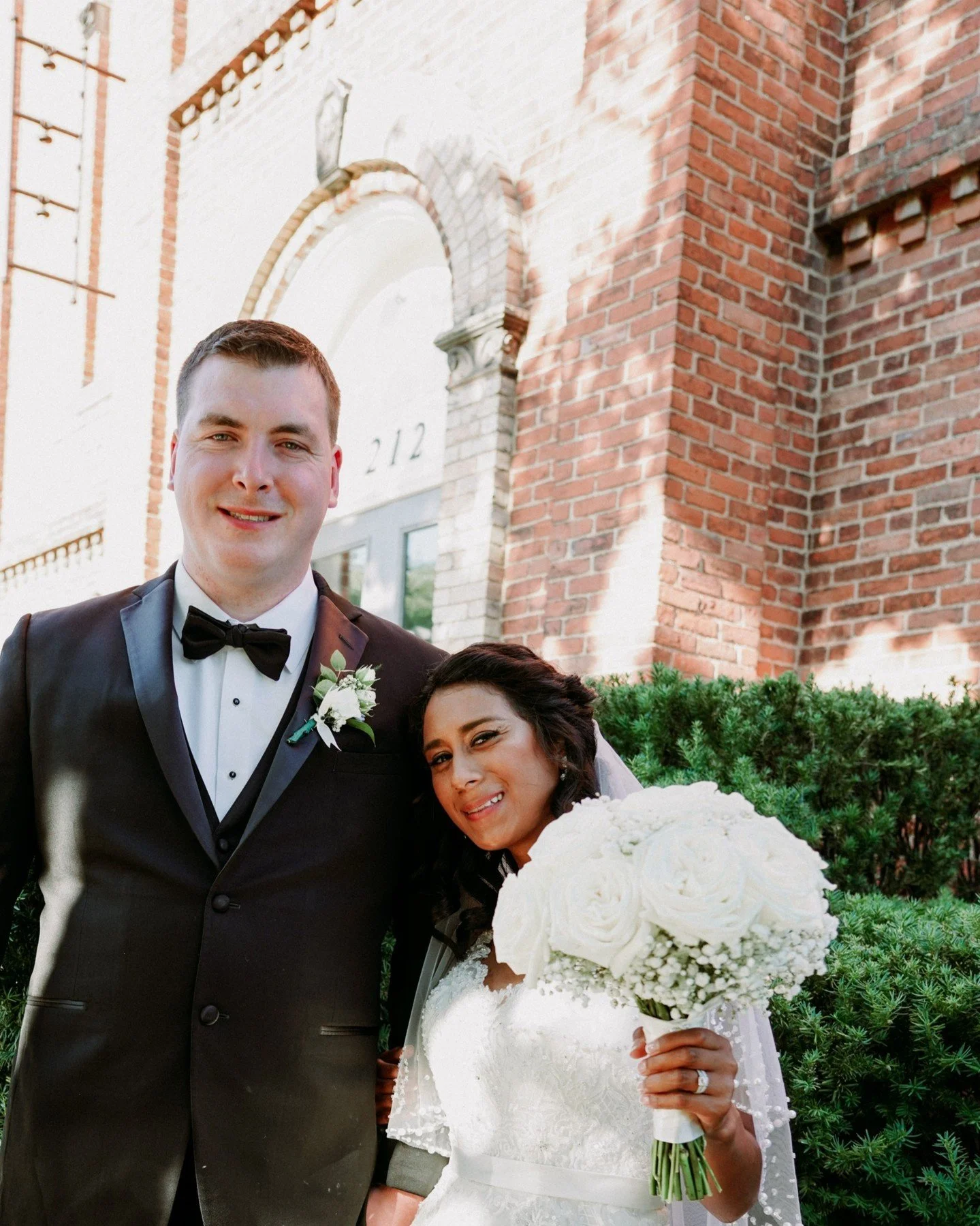 A newlywed couple standing outside a brick building. The groom is wearing a black tuxedo with a smile, and the bride is holding a large white bouquet, smiling, in a white wedding dress.