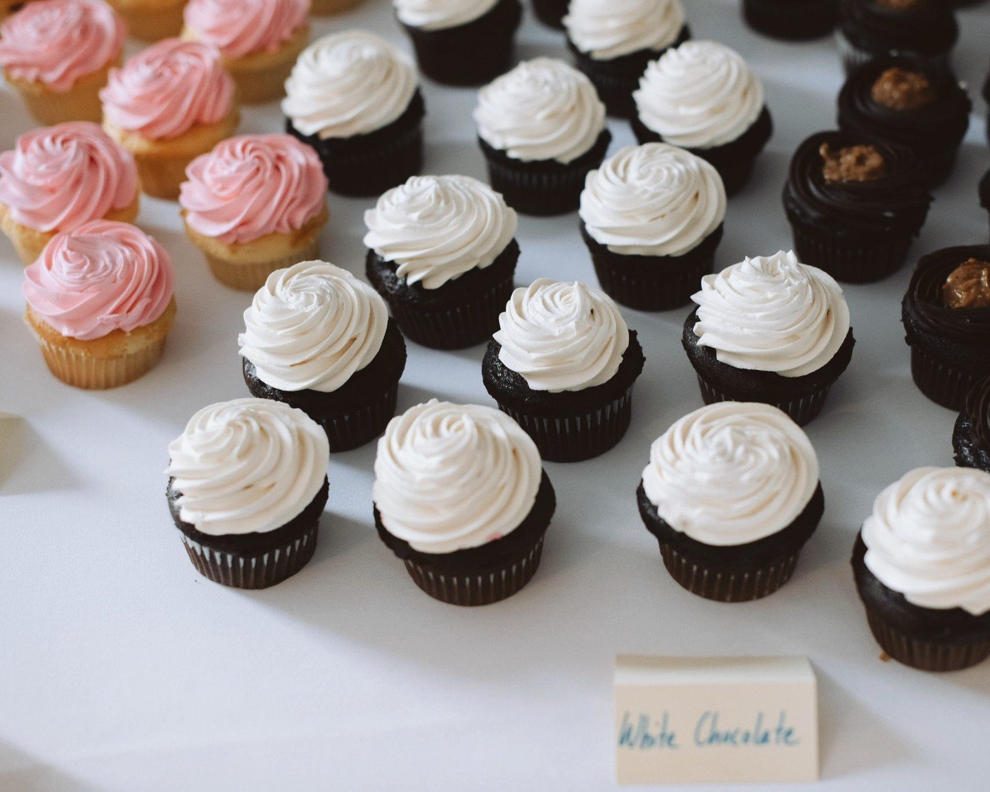 Assorted cupcakes with pink, white, and chocolate frosting, arranged on a white surface, with a label reading 'White Chocolate' in front.