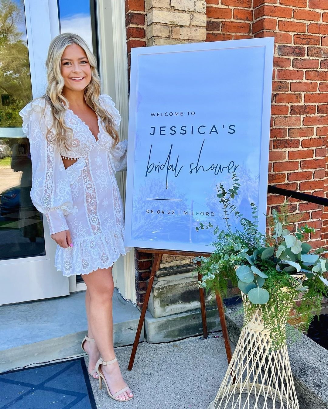 A woman in a white lace dress standing next to a large sign that says "Welcome to Jessica's Bridal Shower 06.04.22 Milford." The woman is smiling and standing outdoors near a brick building, with a decorative flower arrangement in front of the sign.