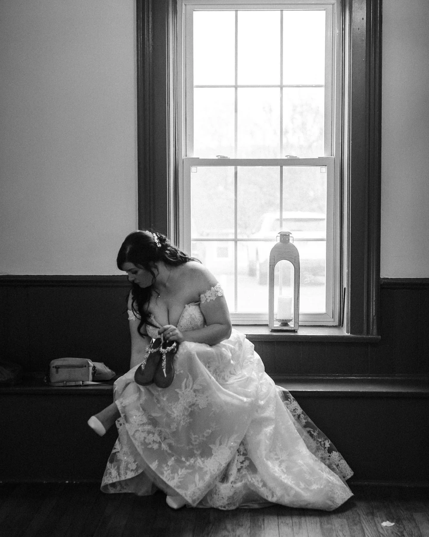 A bride sitting on a wooden bench in a room with a large window, holding her shoes and necklace, with a purse beside her.