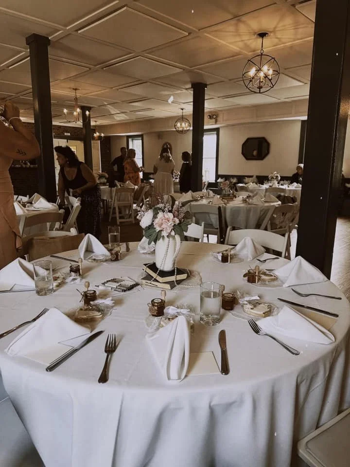 Round banquet table with white tablecloth, decorated with a floral centerpiece in a white vase, surrounded by white chairs. The table is set with white napkins folded into triangles, silverware, glasses, and small jars or candles, in an elegant dining room with other tables and guests in the background.