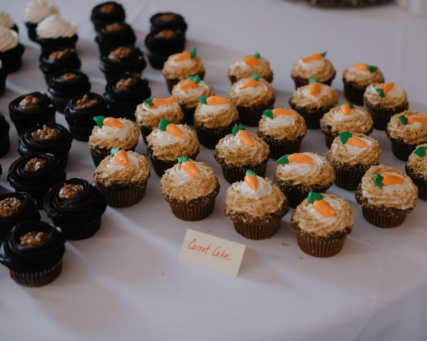 Mini carrot cake cupcakes with cream cheese frosting and small fondant carrots on top, arranged on a white surface with a small sign that reads 'Carrot Cake'.