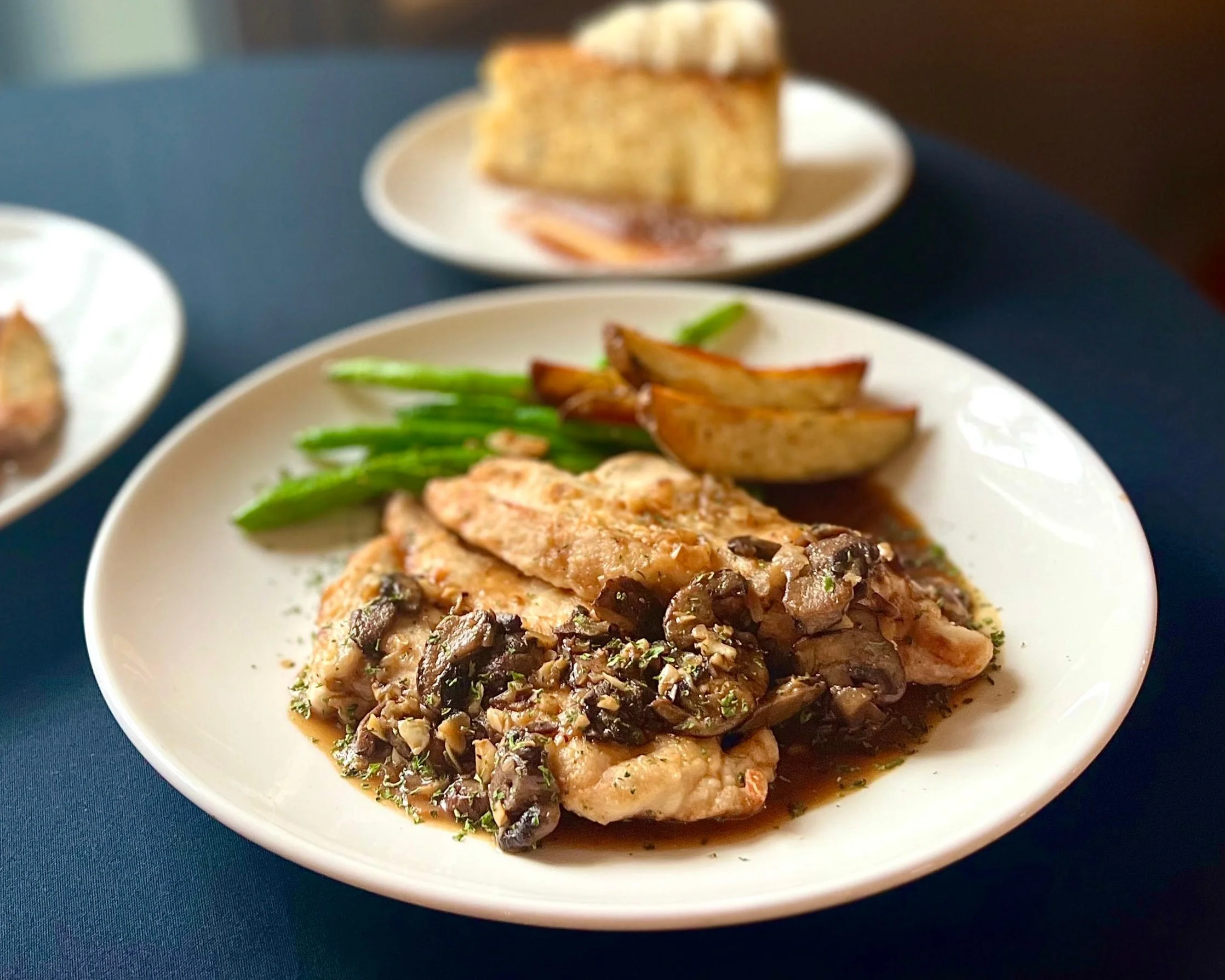 A white plate with cooked chicken topped with mushroom sauce, green beans, and roasted apple slices, with another plate of food slightly out of focus in the background.
