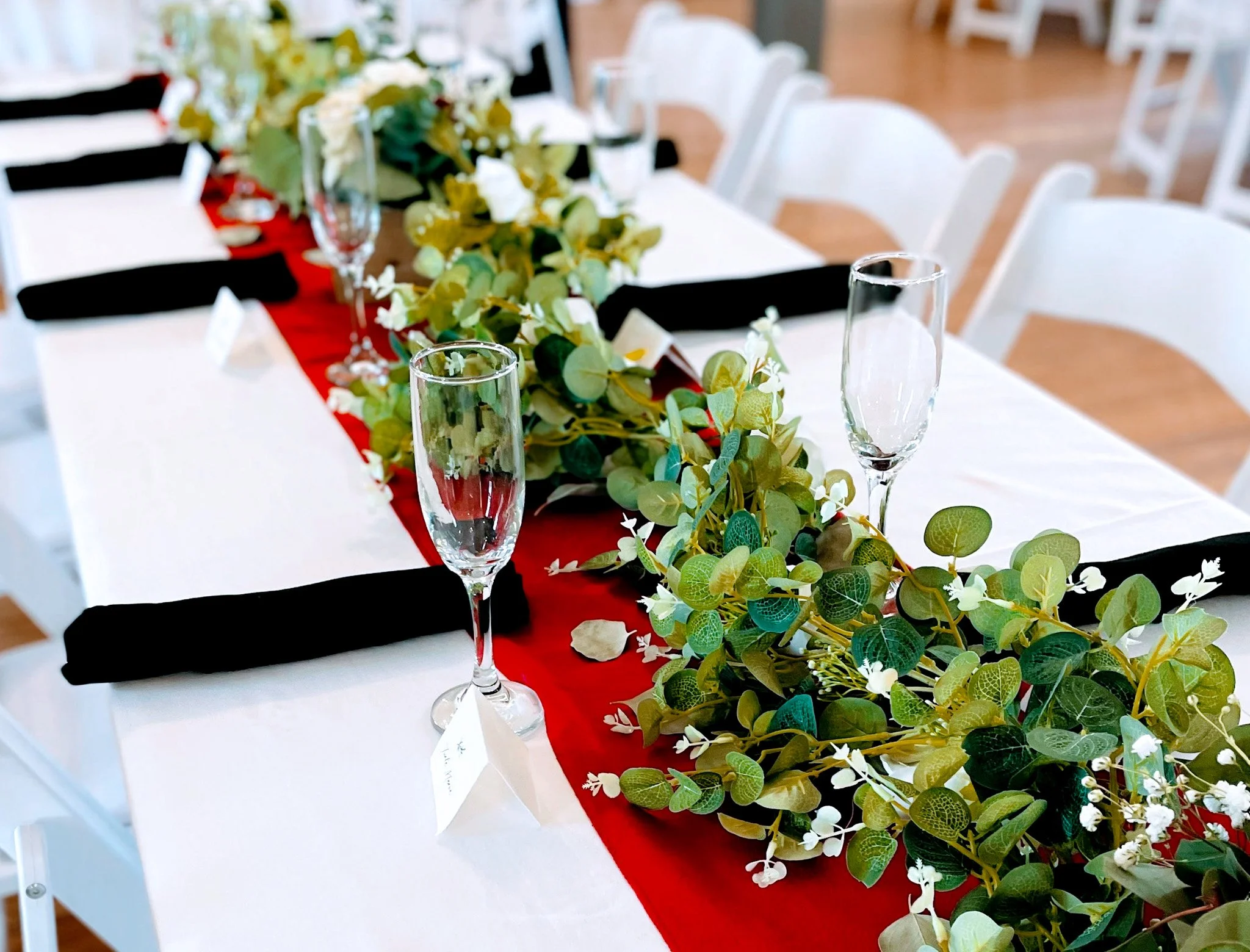 Table decorated with a red table runner, a green leafy garland, white flowers, and empty champagne flutes, set for a celebration or event in a well-lit room.