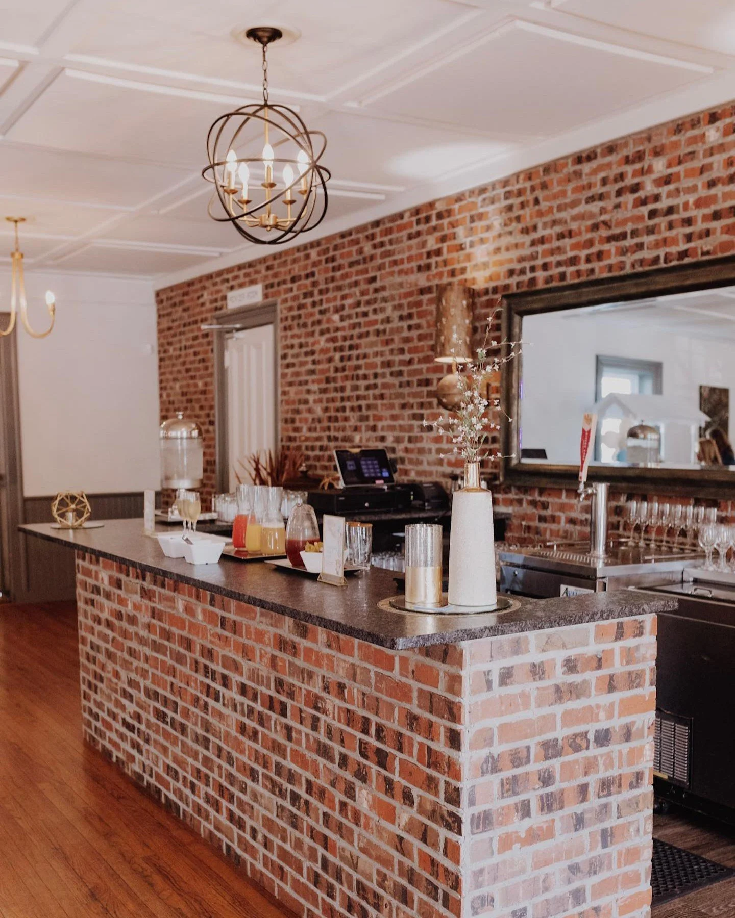 Interior of a restaurant with a brick counter, wooden floors, and a decorated brick wall. Chandeliers hang from the ceiling. The counter has beverage pitchers, glasses, and decorative vases.