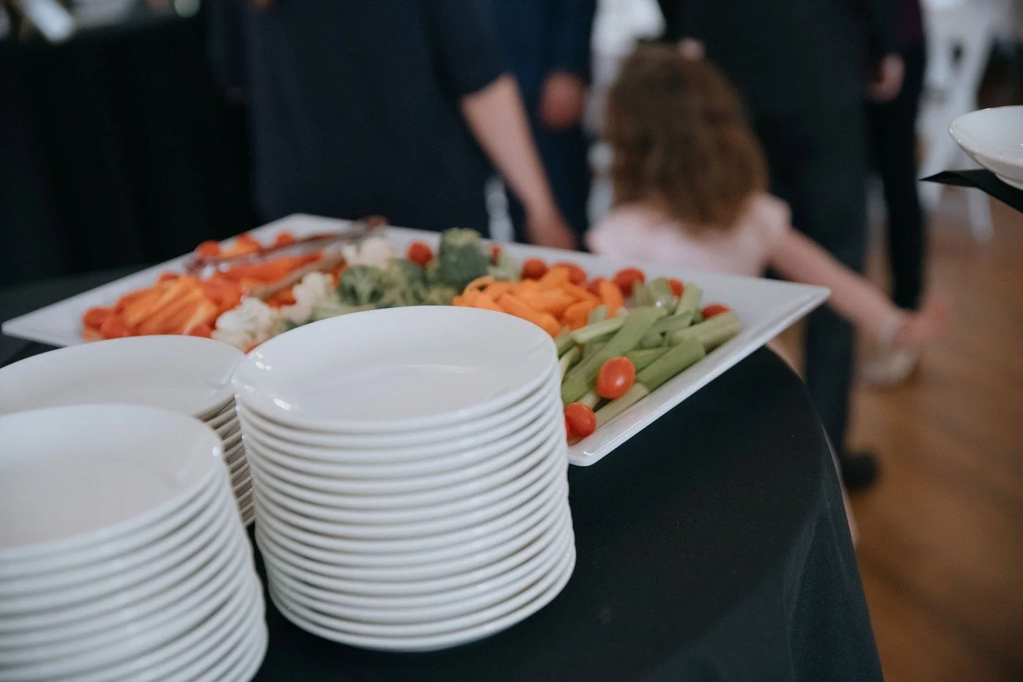 A rectangular white platter with assorted fresh vegetables, including cherry tomatoes, broccoli, carrots, celery, and cauliflower, placed on a black tablecloth. In front of the platter are stacked white bowls, and in the background, blurred people ar