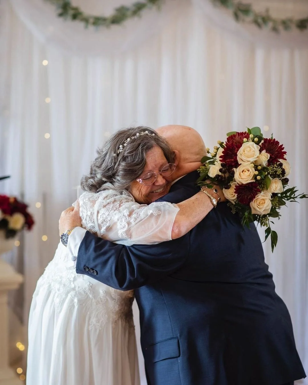 An elderly woman in a white lace dress and glasses hugging a bald man in a dark suit, holding a bouquet of red and white flowers during a wedding celebration.
