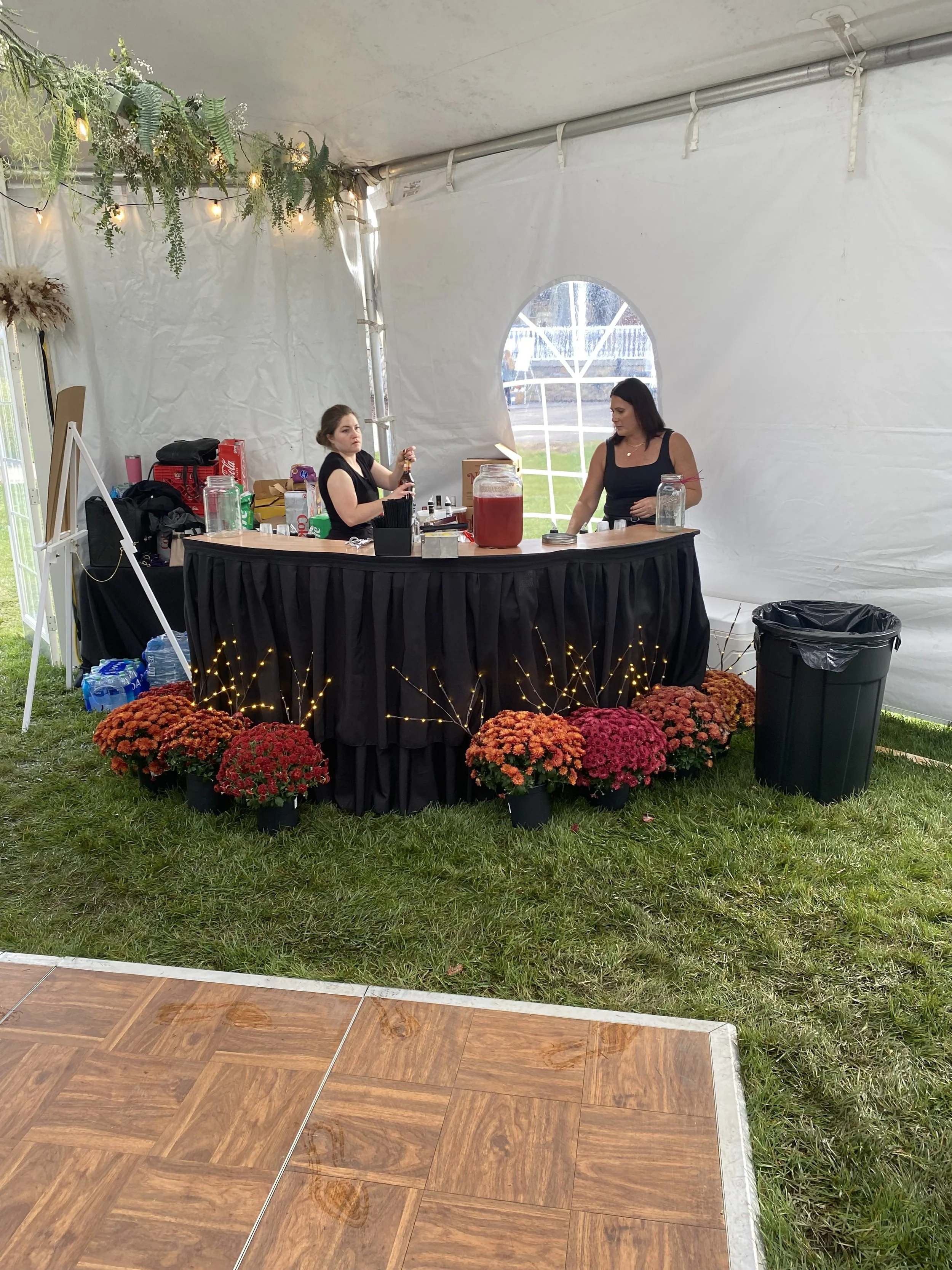 Two women stand behind a black table with various drinks and supplies, inside a white tent decorated with greenery and string lights, with potted orange and red chrysanthemums at the front.