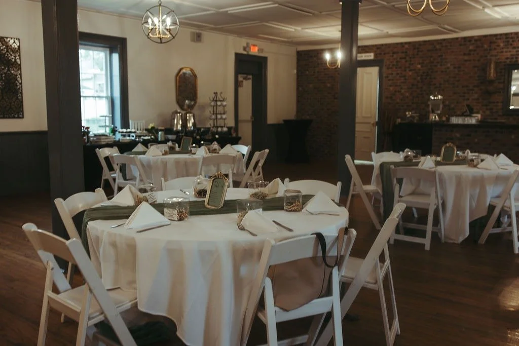 Wedding reception area with round tables decorated with white tablecloths, napkins, and small centerpieces, set inside a rustic venue with brick and wood walls and soft lighting.