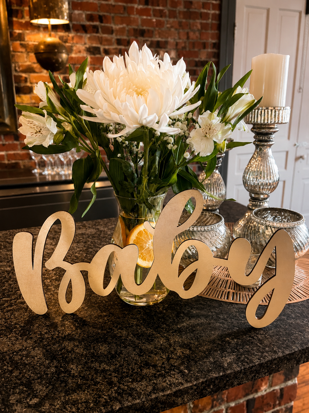 A kitchen countertop decorated with a vase of white flowers, including a large white dahlia and lilies, surrounded by silver candle holders and a gold 'happy' sign. The background features a brick wall and a mirror with wine glasses.
