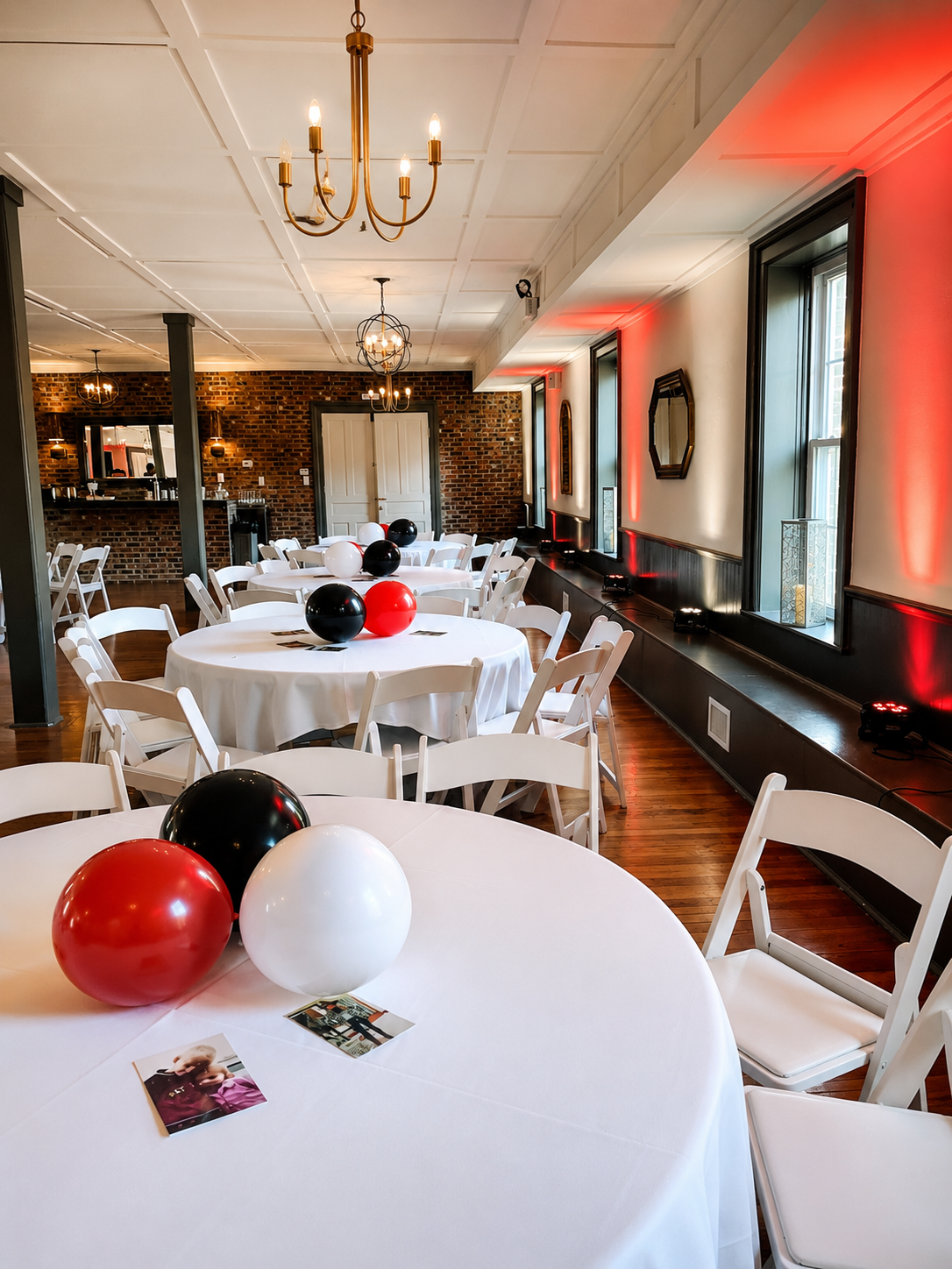 Indoor event space decorated with white round tables, each with black, white, and red balloons, and a few photographs. The room has large windows with black frames, brick and white walls, wooden floors, and elegant chandeliers. Red uplighting accents