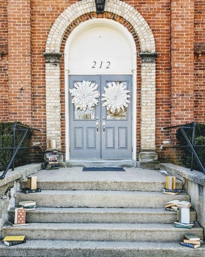 A brick building entrance with a rounded white arch, a gray door decorated with white wreaths, stairs, and books scattered on each step.
