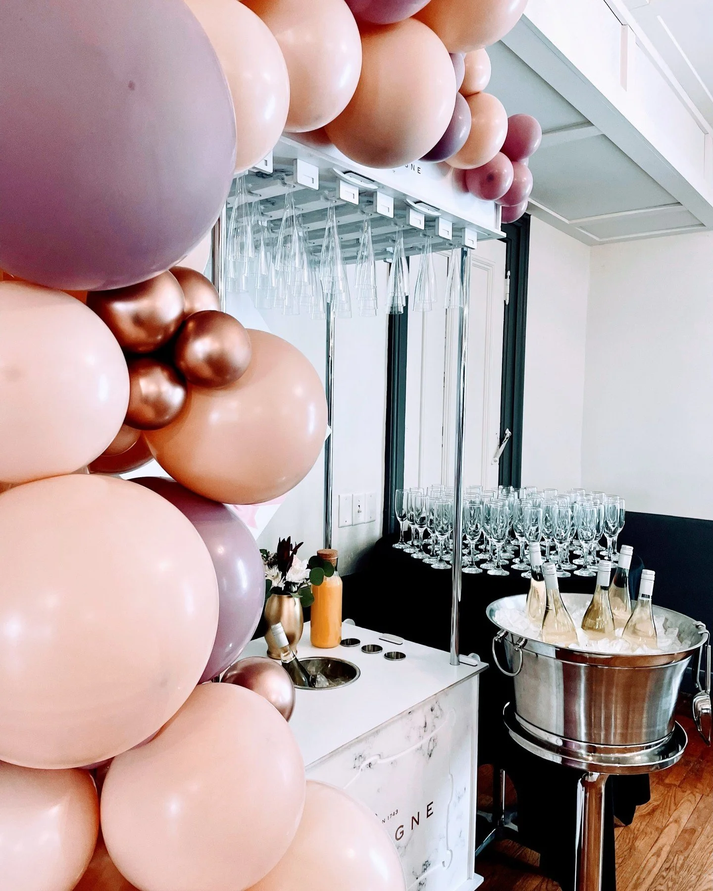 Party setup with pink and rose gold balloons, a table with glassware, champagne bottles in an ice bucket, and a drink station with flowers and utensils, in an indoor venue.