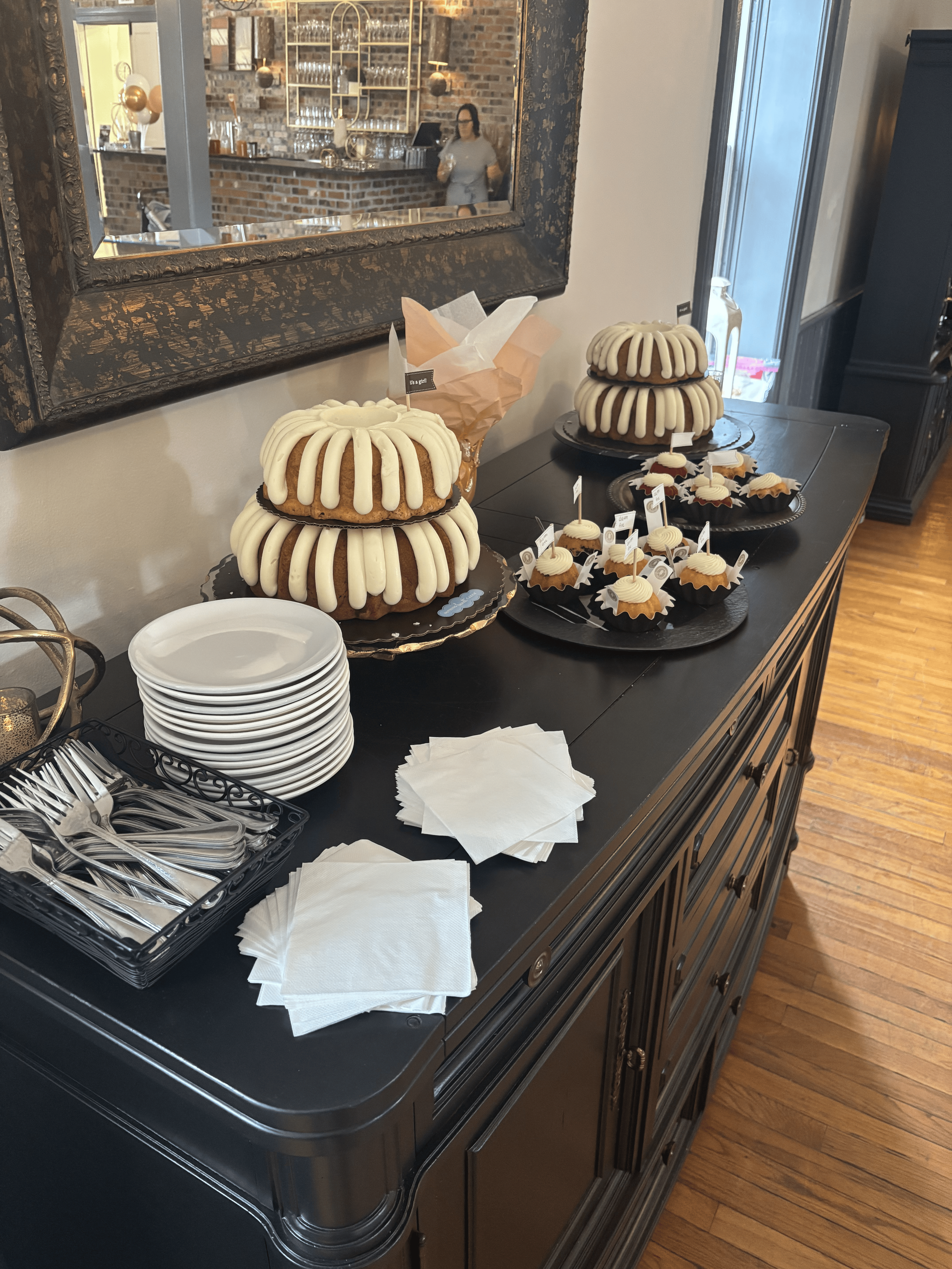 A table with two large Bundt cakes covered with white icing and small cupcakes, all decorated for a celebration, with plates, napkins, and utensils nearby.