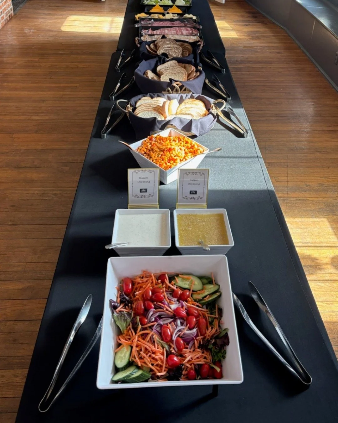 A buffet with a variety of salad toppings and dressings. There is a large bowl of cherry tomato salad, smaller bowls of ranch and Italian dressing, and containers with sliced cucumbers, shredded carrots, and other vegetables. The buffet is set on a l