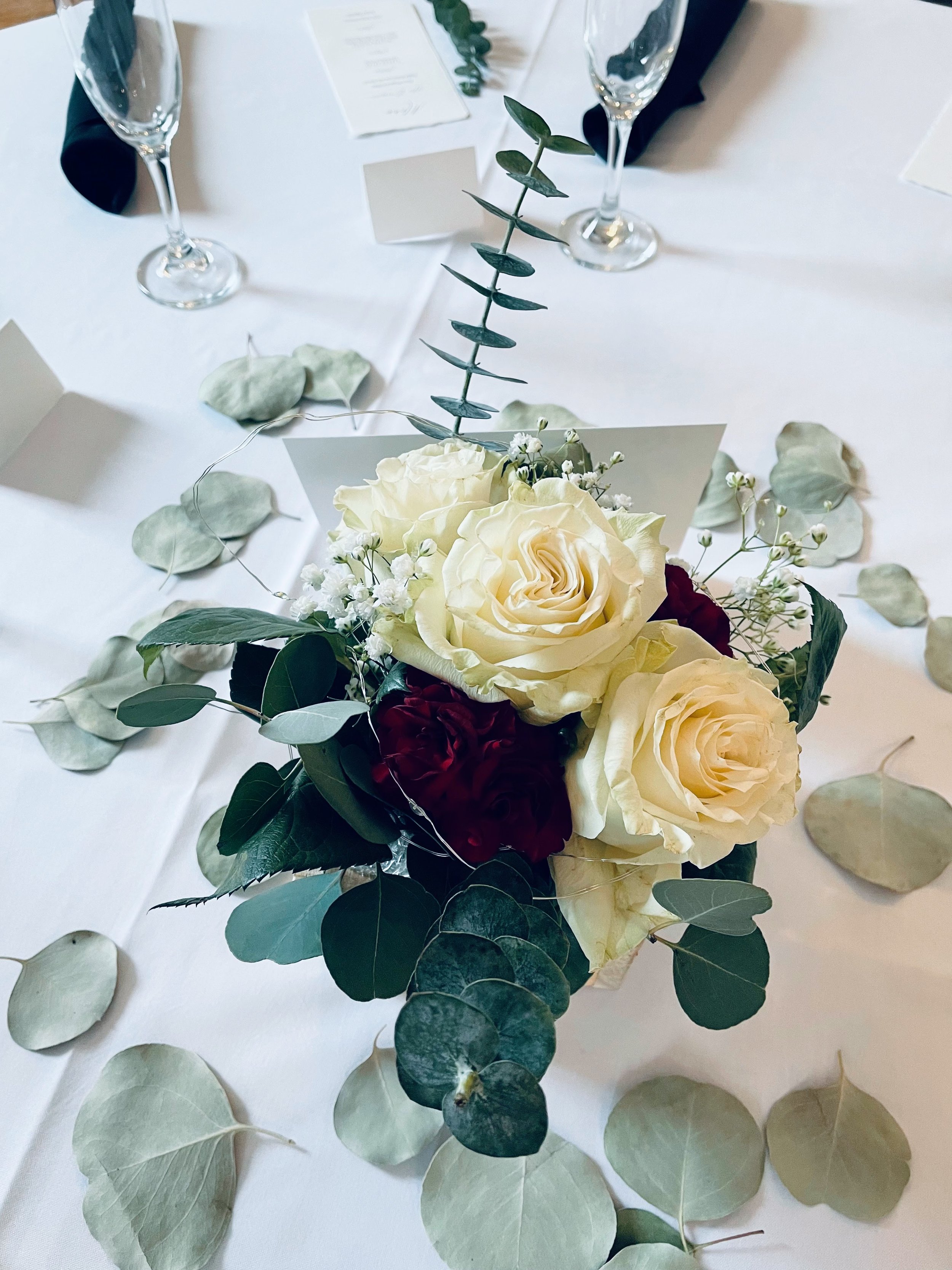 A floral centerpiece with white roses, baby’s breath, eucalyptus leaves, and green foliage on a table with white tablecloth and scattered eucalyptus leaves.
