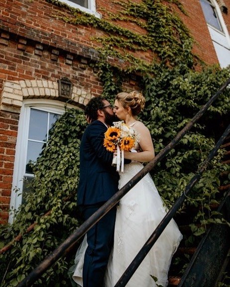 Bride and groom sharing a kiss outdoors on a staircase surrounded by green ivy, with a brick building in the background, during their wedding wedding.