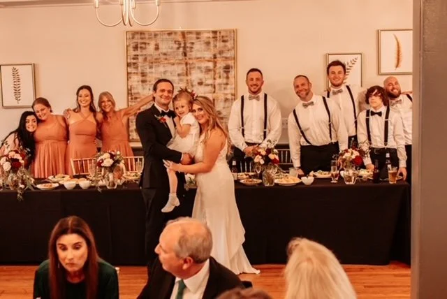 Group of people at a wedding reception, with a bride and groom in front, surrounded by family and friends, seated at a reception table with floral centerpieces.