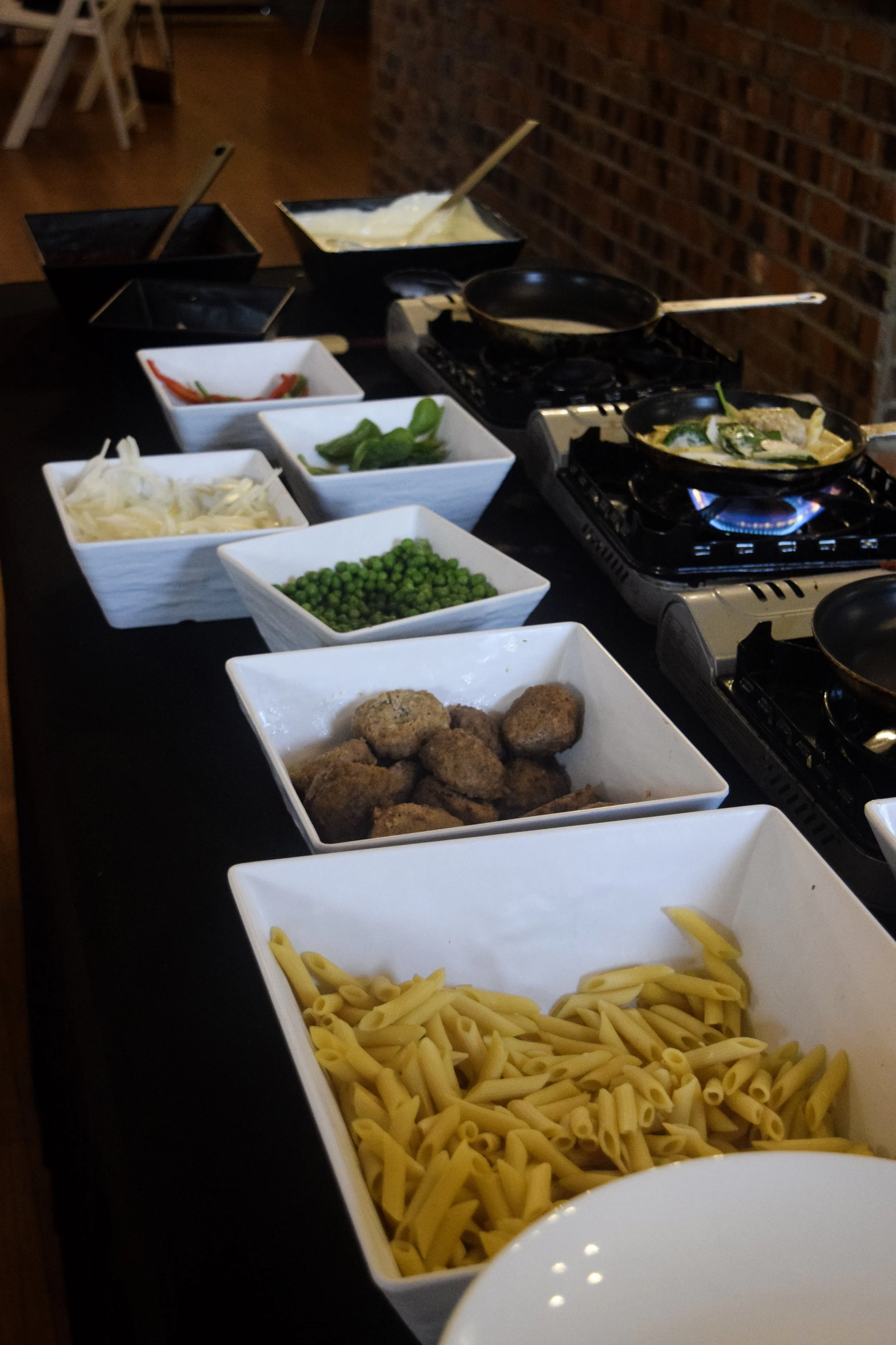 A countertop with various ingredients for cooking, including bowls of pasta, peas, shredded cheese, and meatballs, alongside two stovetop burners with pans in use.