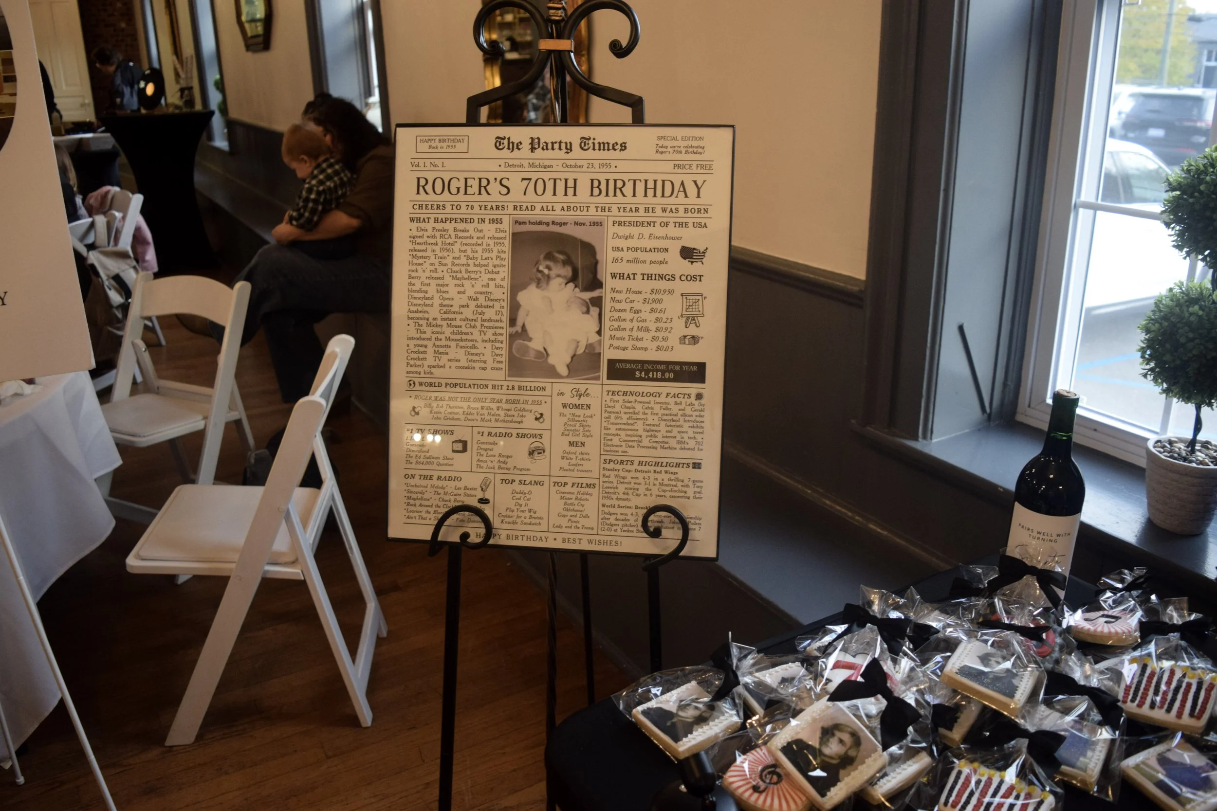 A vintage newspaper-style display board commemorating Roger's 70th birthday, with a photo and historical facts, set in a room with white chairs, a window, and some gifts and decorations visible.