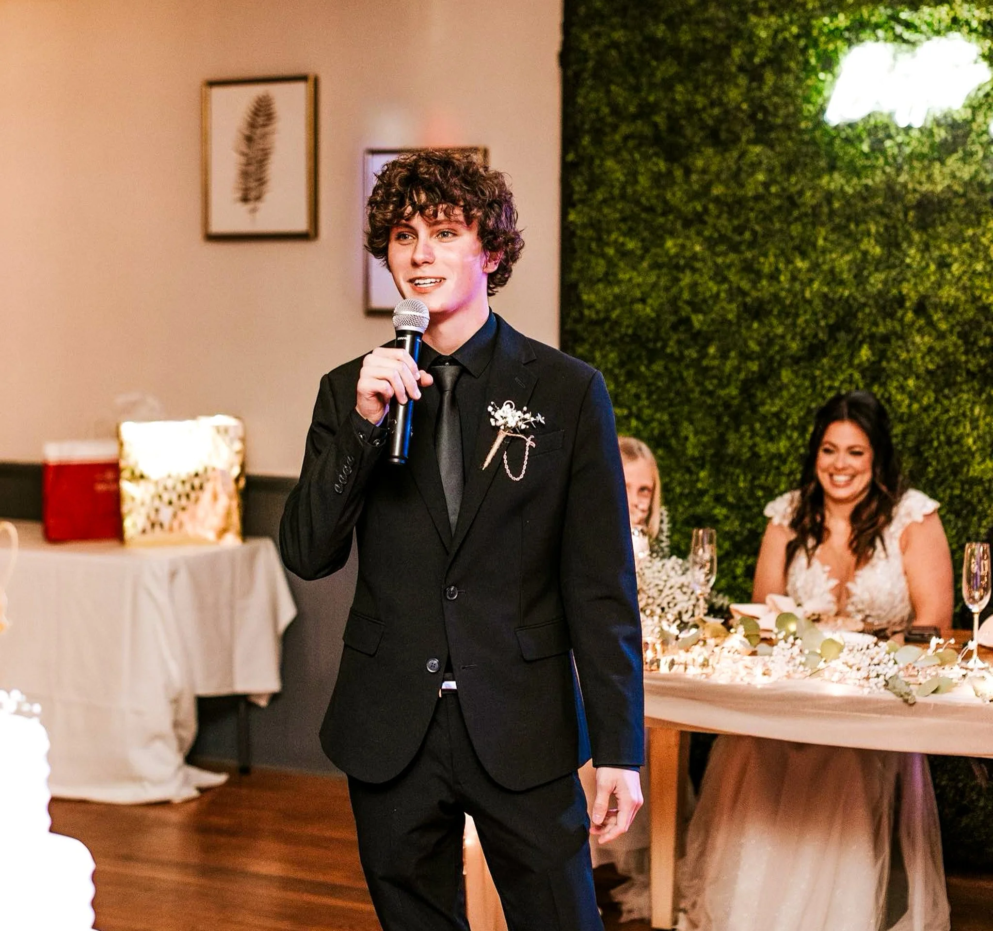 A young person with curly hair wearing a black suit and holding a microphone, speaking at a formal event decorated with a green leafy wall and framed art, with guests seated at a table.