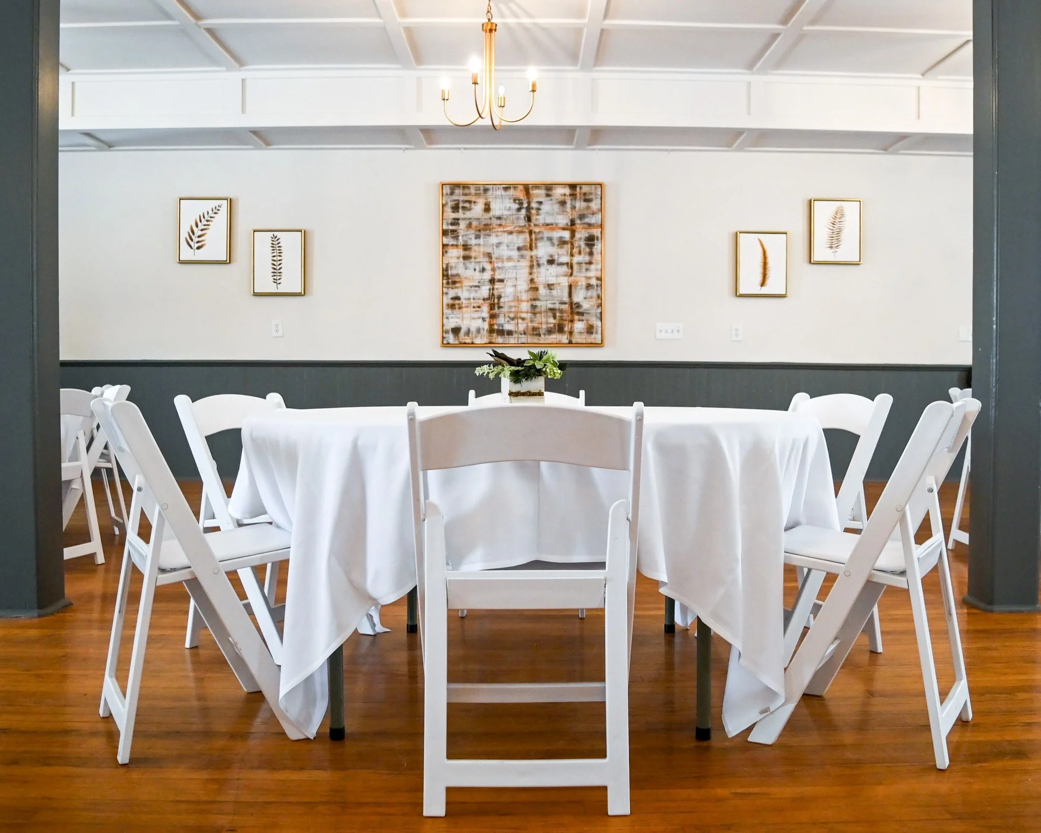 A dining room with a round table covered with a white tablecloth, surrounded by white folding chairs, and a floral centerpiece. The wall behind features framed botanical prints and an abstract painting, with a chandelier hanging from the ceiling.