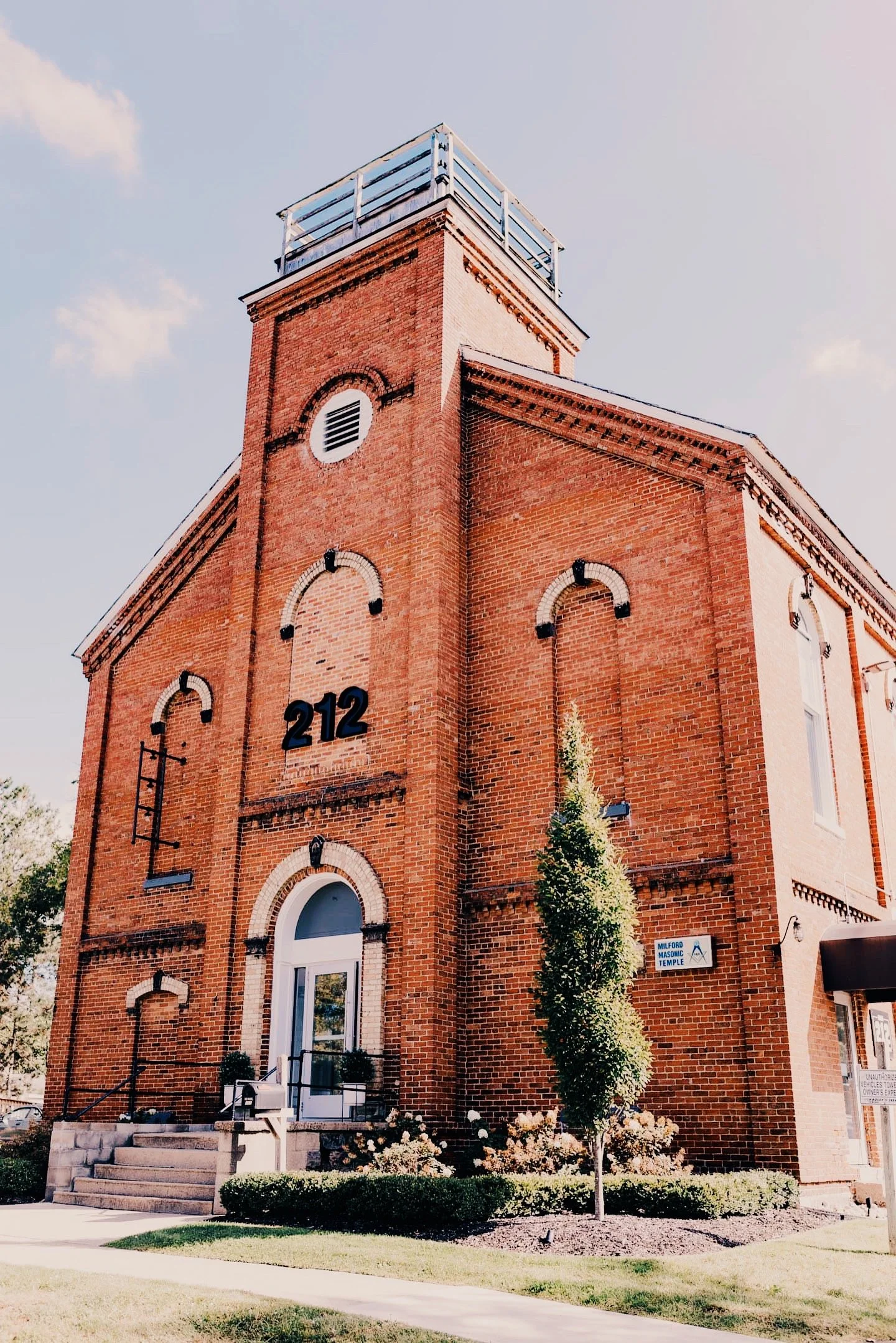 Red brick building with the number 212 on it, labeled as Milwaukee Masonic Temple, with steps leading up to the entrance, surrounded by greenery and flowers, under a partly cloudy sky.