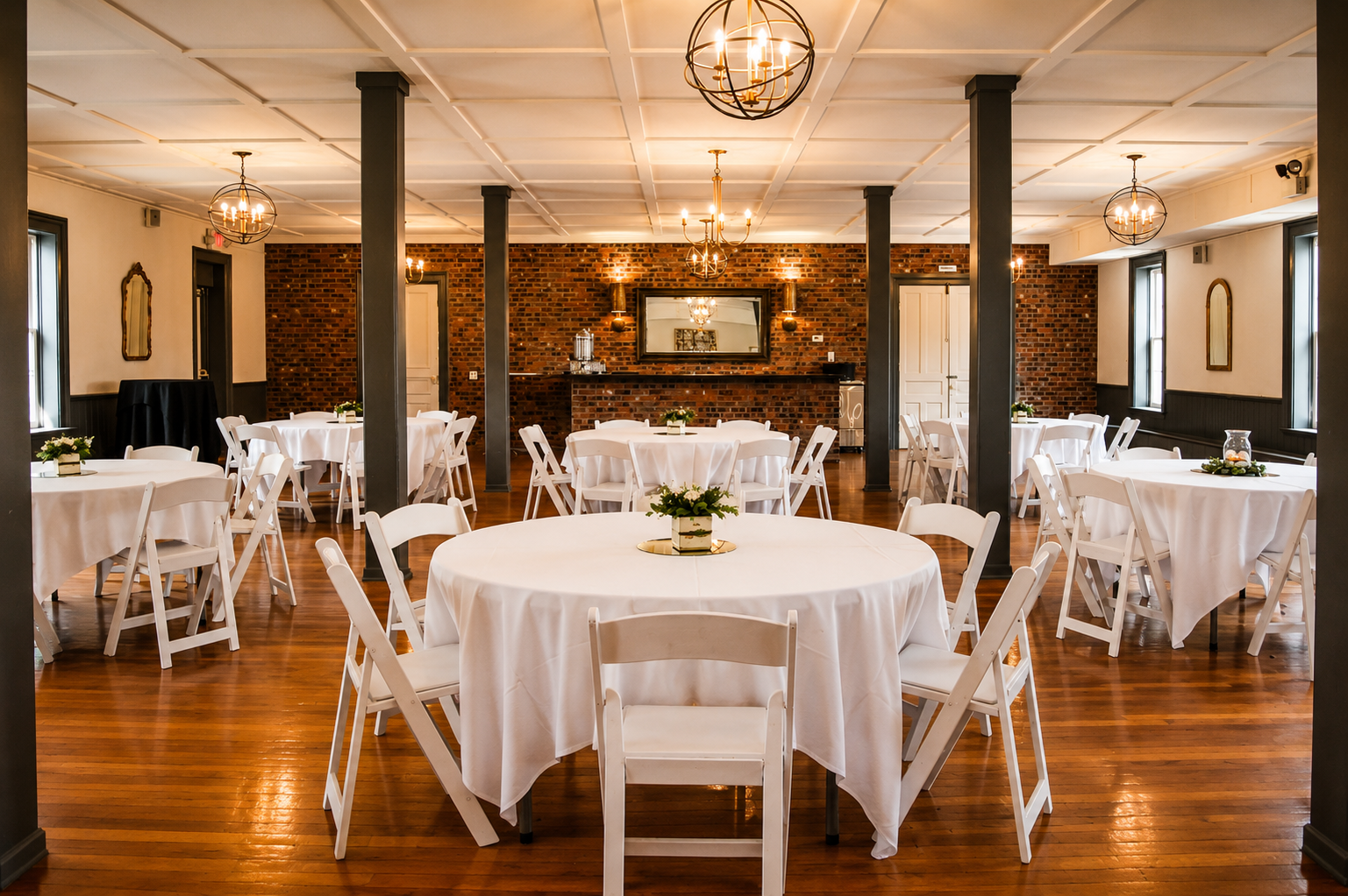 Elegant banquet room with white round tables and chairs, decorated with small floral centerpieces, a brick wall with a large mirror, and multiple chandeliers hanging from the ceiling.