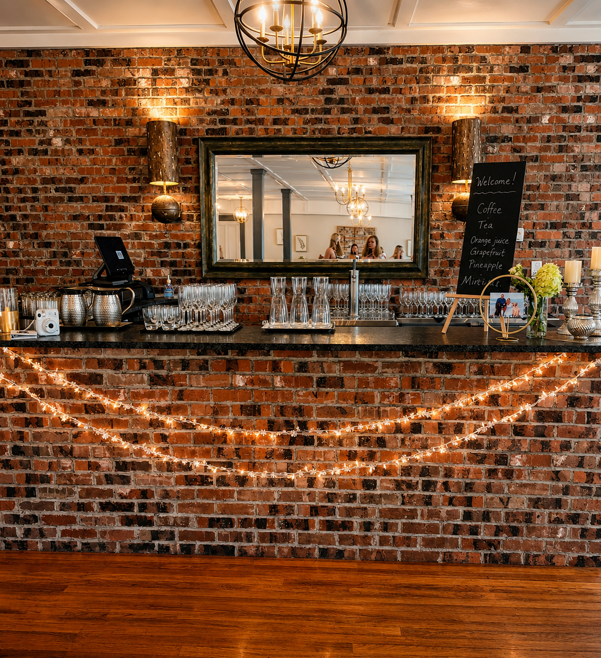A brick bar area decorated with string lights, a large mirror, and a chalkboard menu listing coffee and juice options, with glasses and bar accessories on the counter and chandeliers visible in the mirror's reflection.