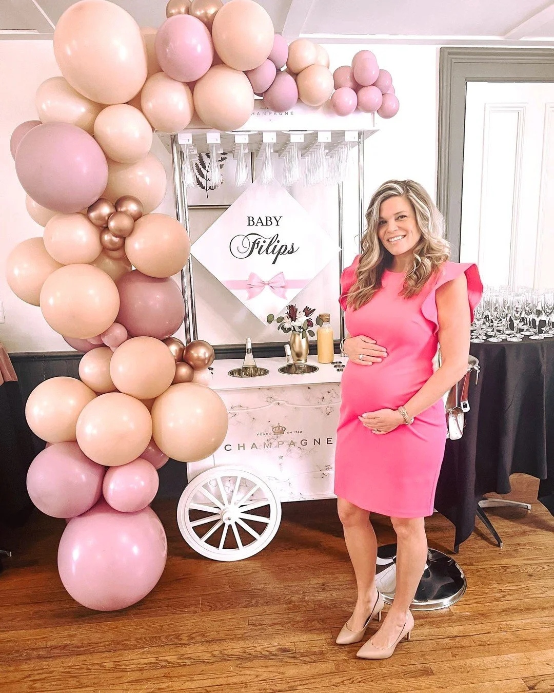 Pregnant woman in a pink dress standing next to a baby shower decoration with pink and gold balloons and a sign that says "Baby Filips" in a decorated indoor space.