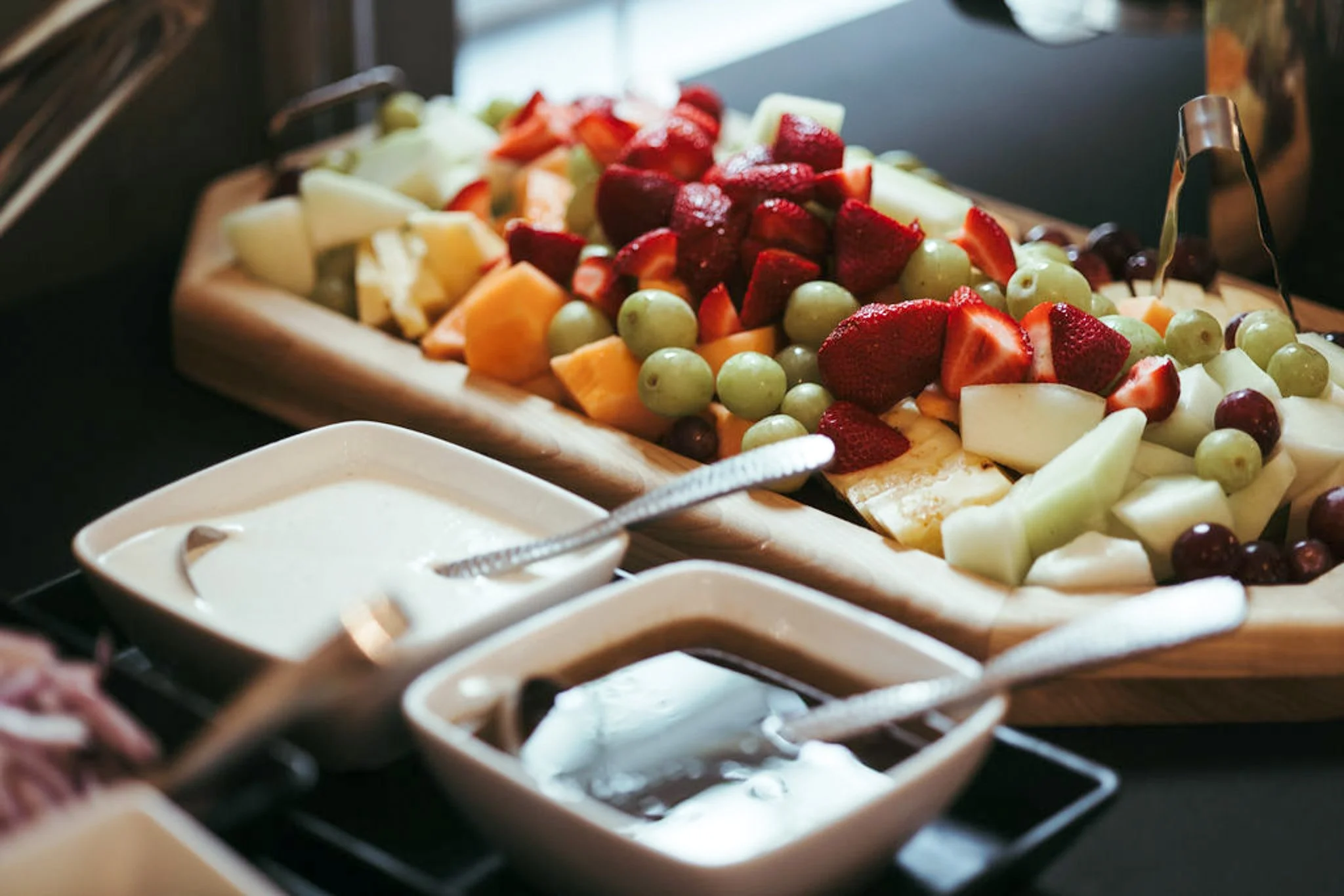 Assorted fresh cut fruits including strawberries, grapes, cantaloupe, honeydew, and pineapple on a wooden platter, with small bowls of dipping sauces in front.