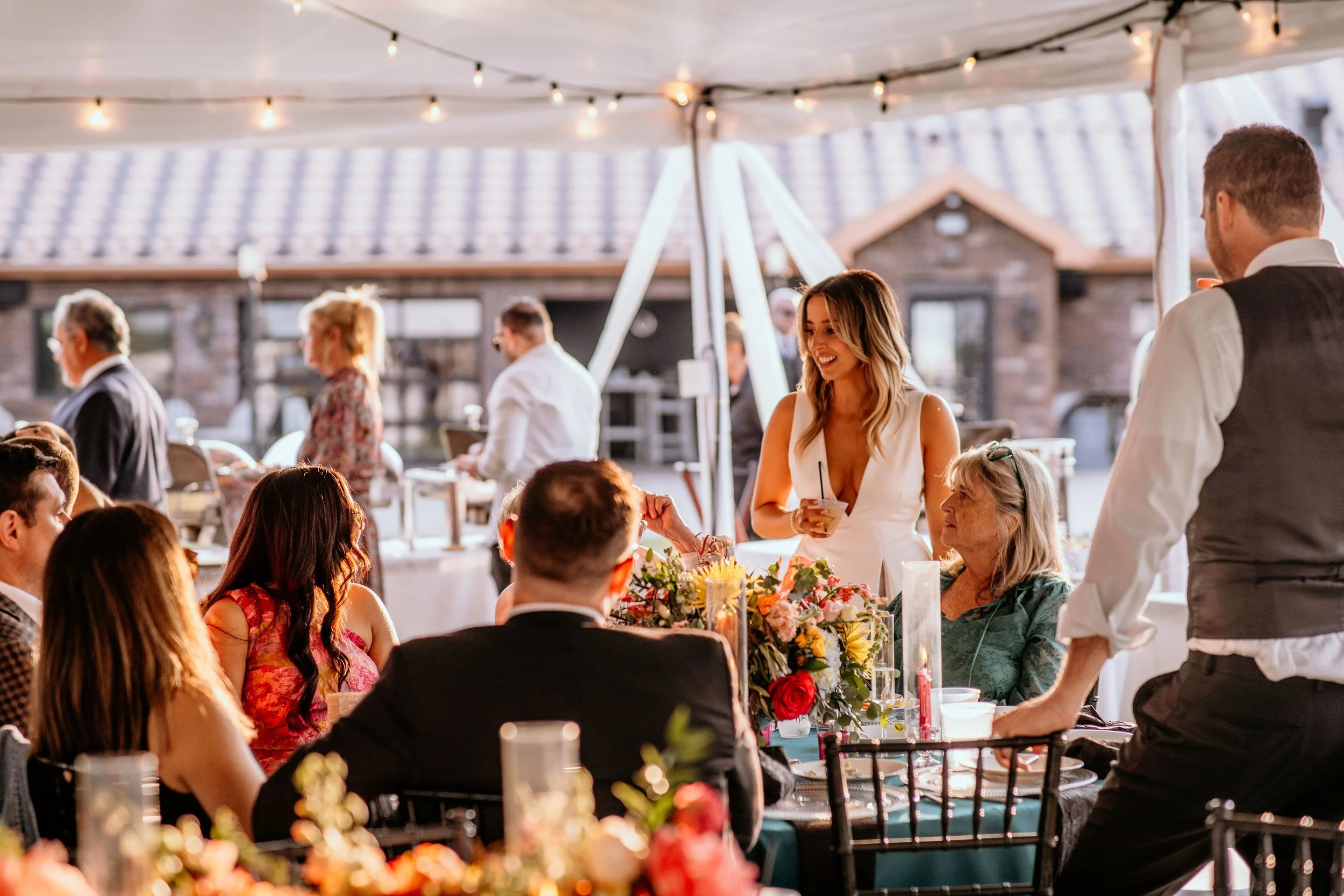 People enjoying a social gathering or celebration under a canopy with string lights, with a woman in a white dress smiling and holding a drink, and others seated at tables with floral centerpieces.