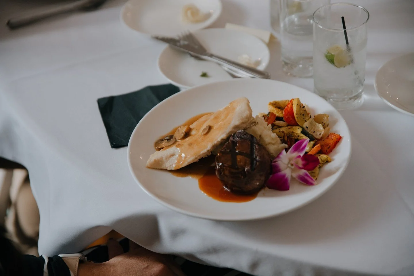 Plate of food with mashed potatoes, beef tenderloin with gravy, and roasted vegetables, garnished with an orchid flower, on a white tablecloth with drinks and utensils.