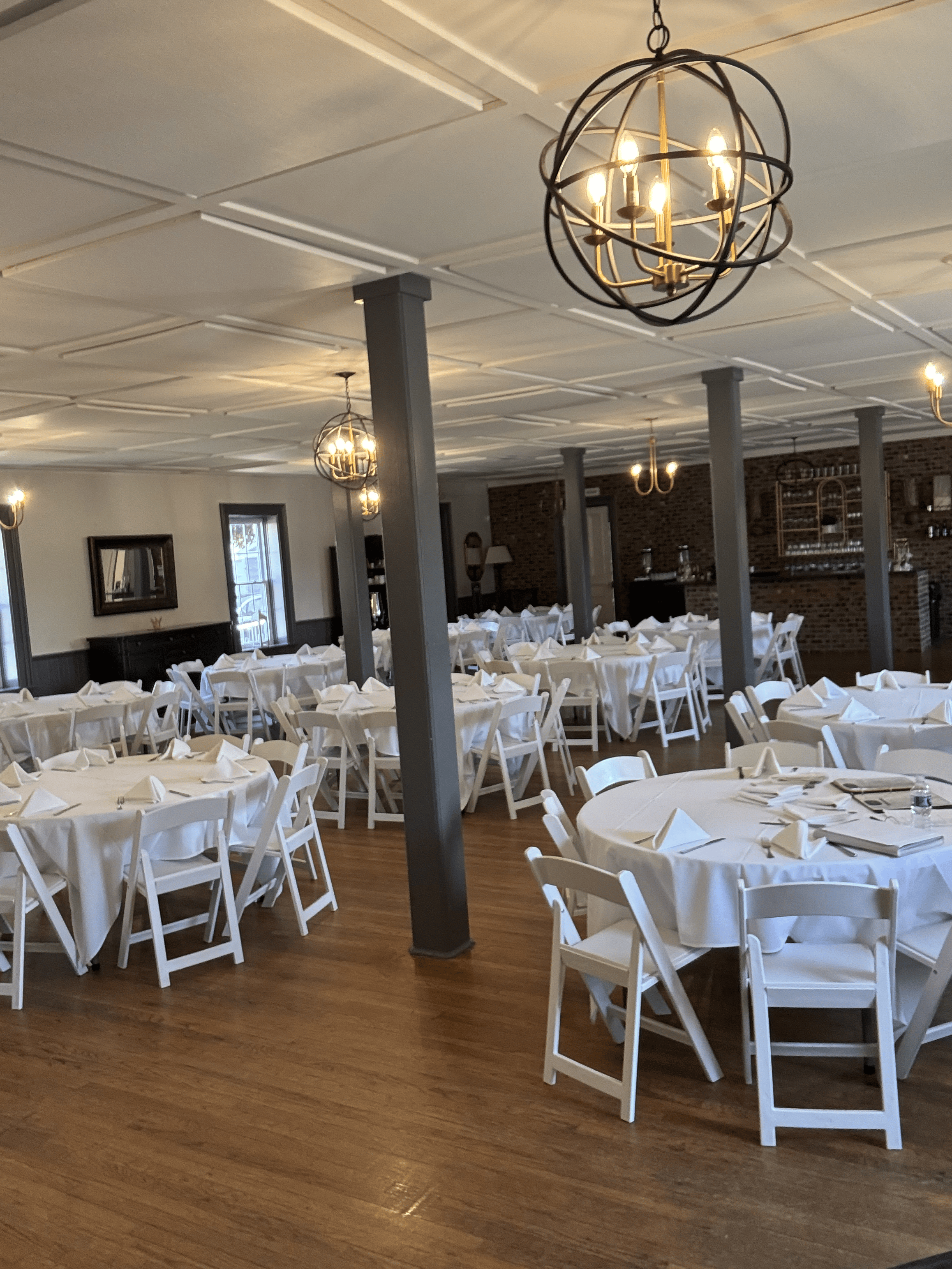 Event space with round tables covered in white tablecloths, white folding chairs, hanging chandeliers and ceiling lighting, wooden floor, brick wall in background, windows, and decorative mirror.