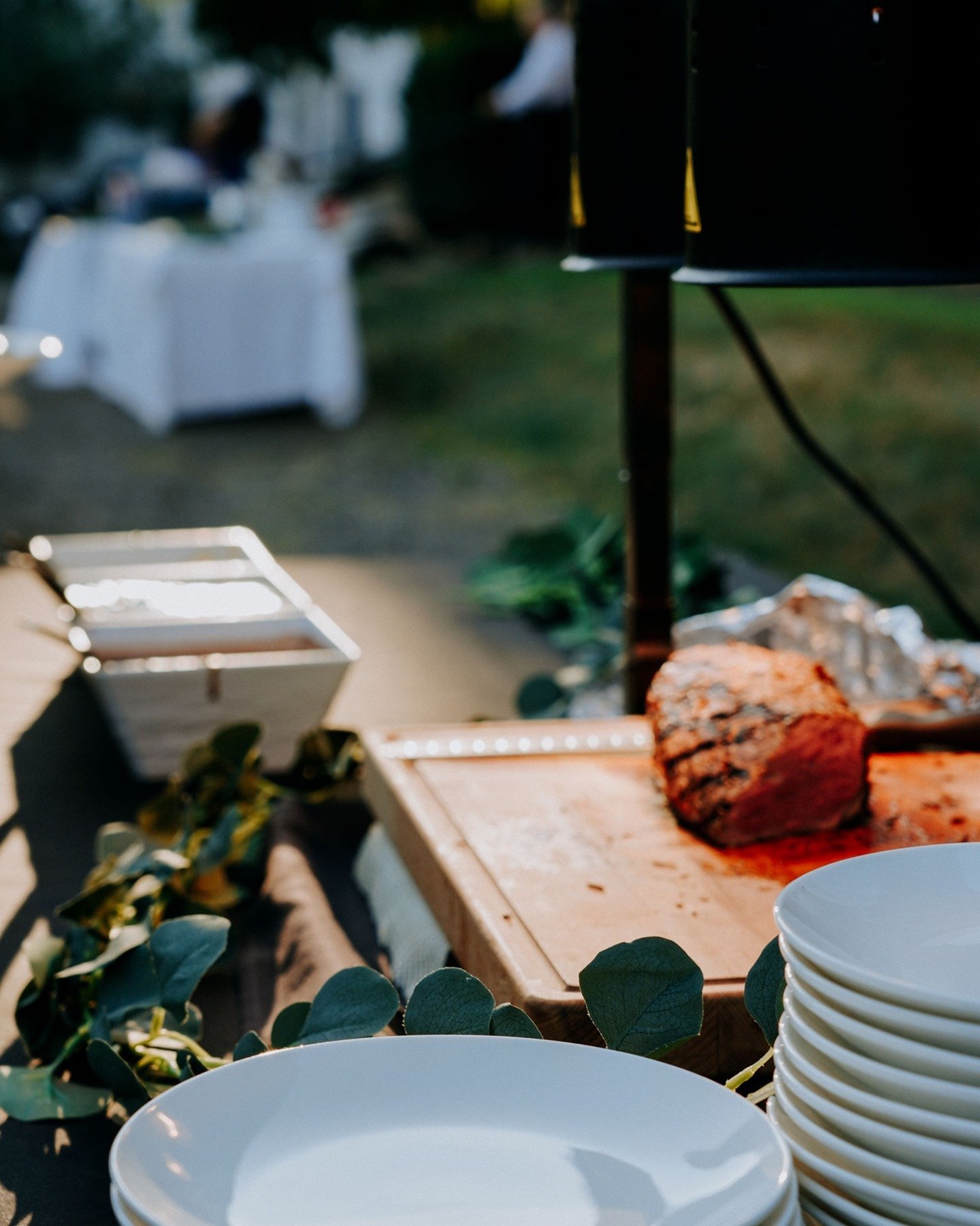 Close-up of white bowls stacked on a table in the foreground, with a barbecue grill and cooked steak in the background at an outdoor gathering.
