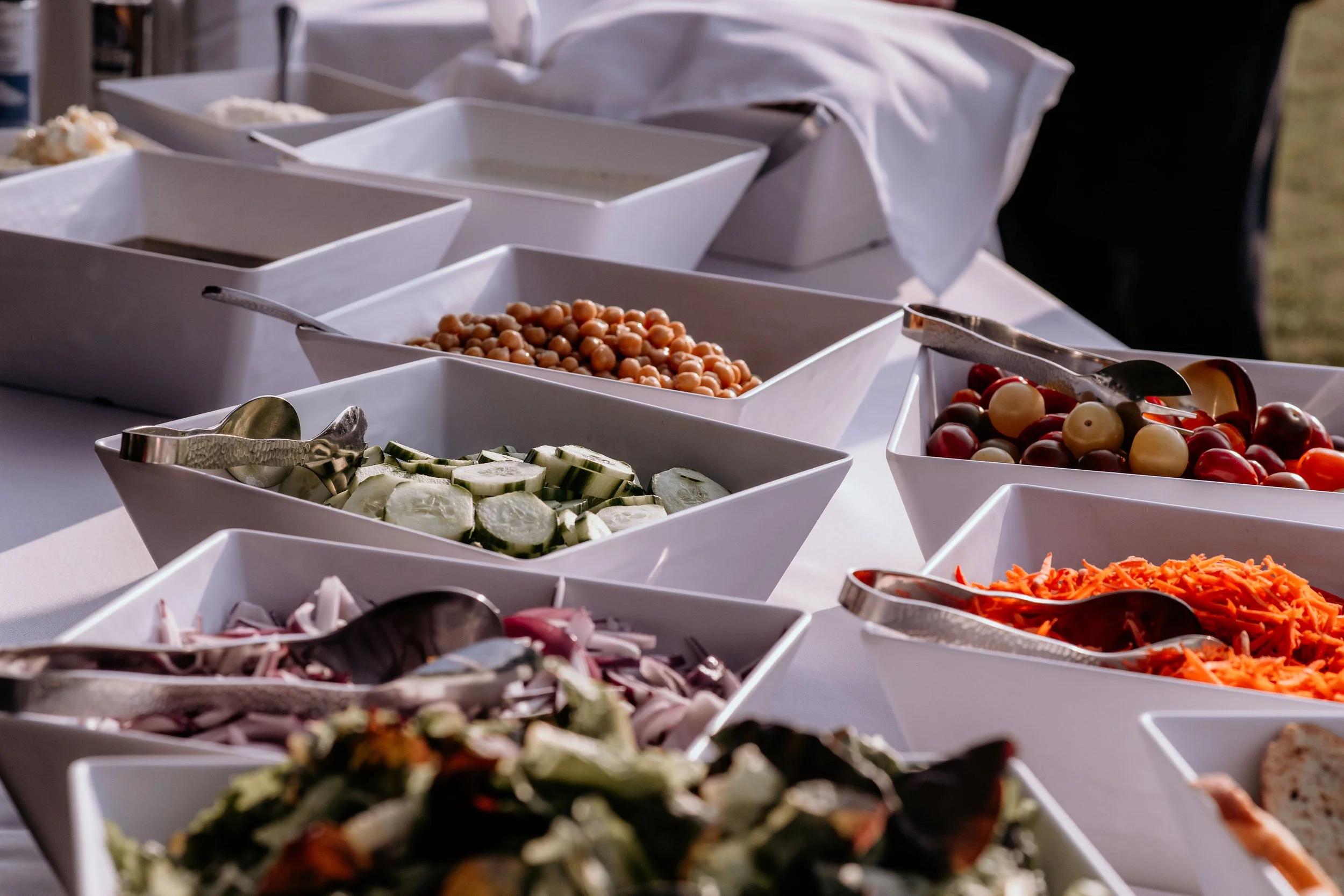 A buffet table filled with white bowls of various salads and toppings, including cucumber slices, chickpeas, grapes, and shredded carrots, with serving spoons in each bowl.
