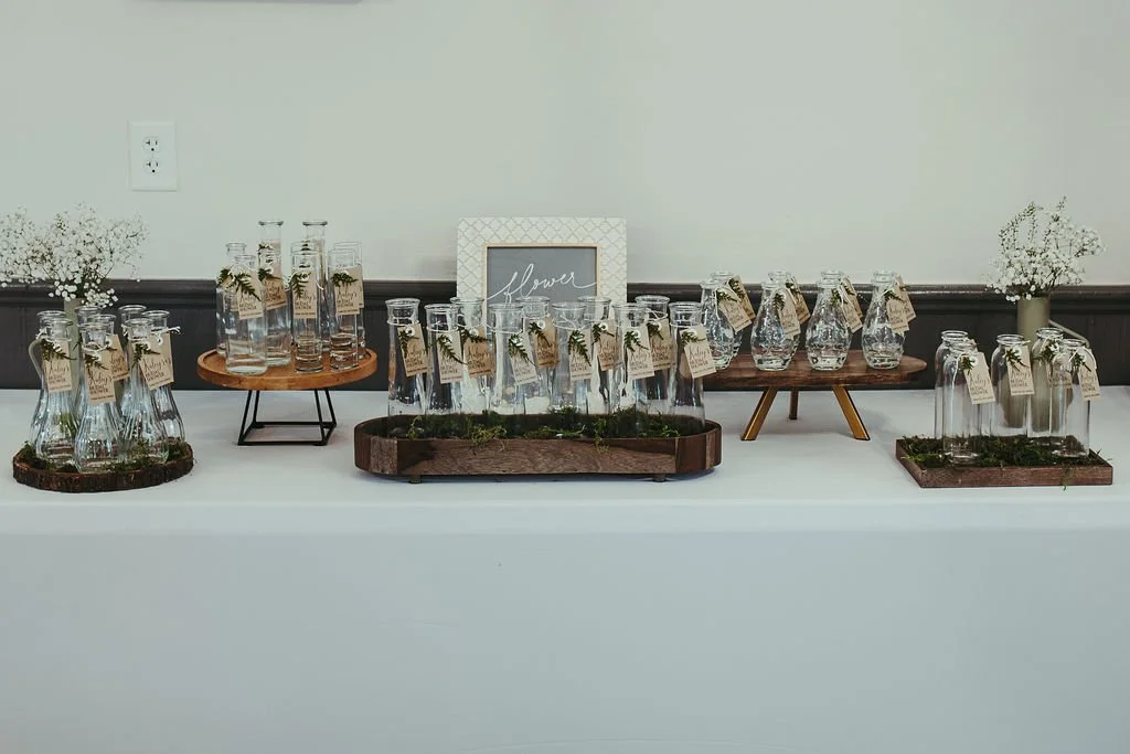 Glass jars and vases with small white flowers, arranged on trays and stands on a white table.