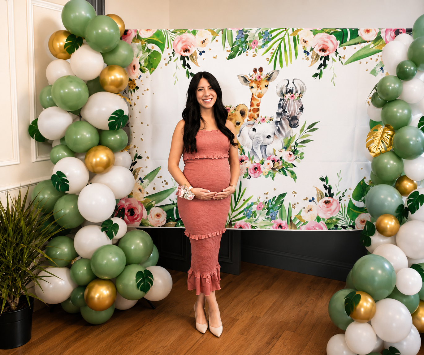A pregnant woman in a pink dress, smiling and holding her belly, standing in front of a colorful jungle animals and floral backdrop, surrounded by balloon arch decorations in green, white, and gold with tropical leaf accents.