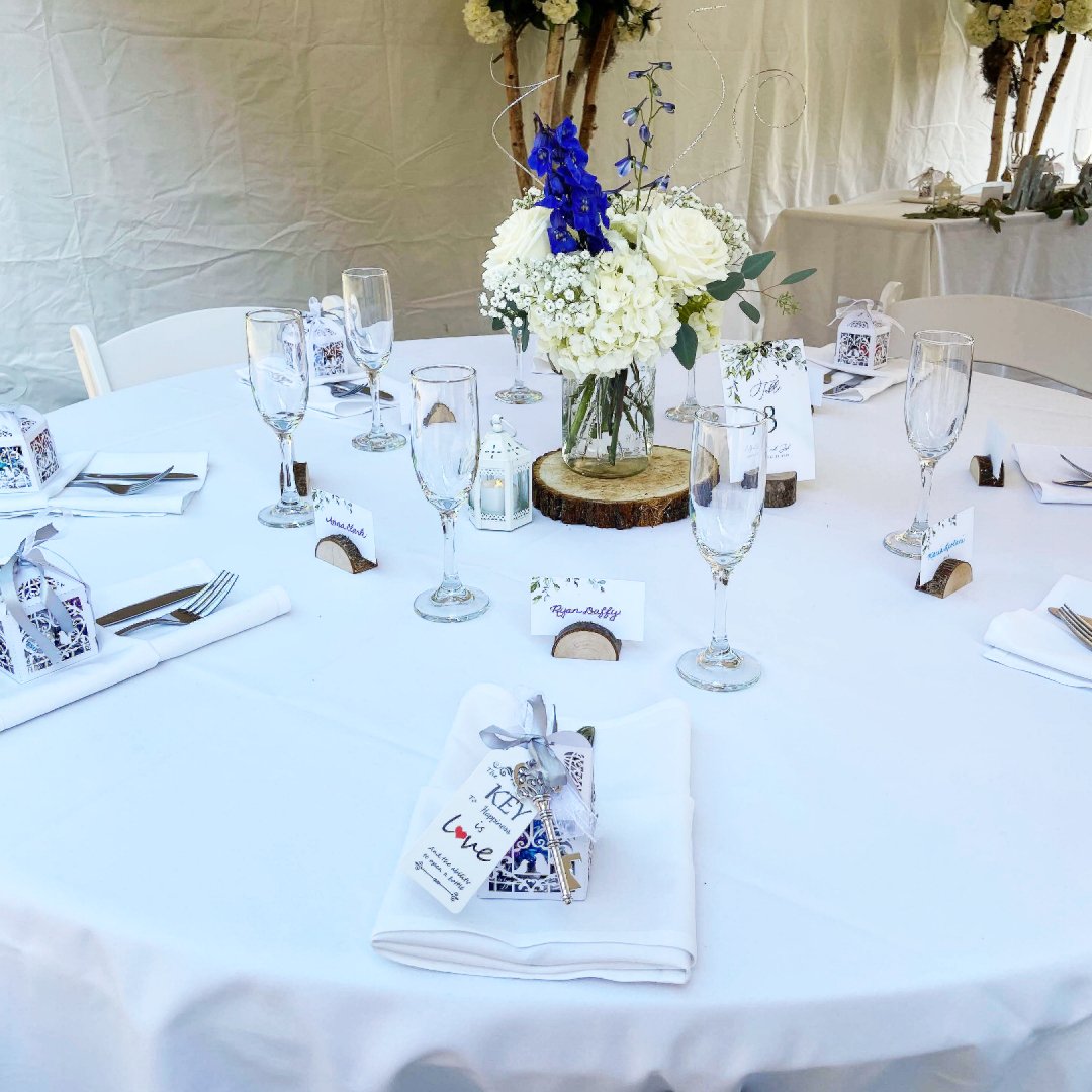 Elegant round table set for a wedding reception with a white tablecloth, featuring a floral centerpiece of white and blue flowers, surrounded by champagne flutes and place settings with wrapped favors and table cards.